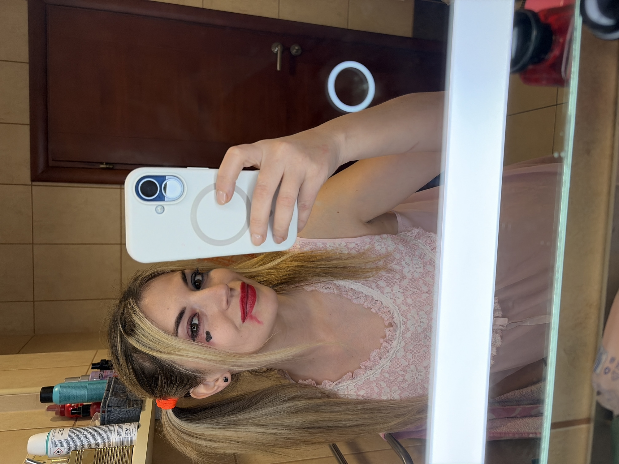 The image is a selfie taken in a bathroom. A woman with blonde hair styled in pigtails is holding a white smartphone up to a mirror, capturing her reflection. She is wearing a pink top with lace detailing. Her makeup includes red lipstick and a heart shape drawn under her eye. In the background, a dark wooden cabinet with a circular light fixture is visible. Various toiletries and hair products are lined up on a shelf next to the mirror. The image does not provide enough information to determine a specific location in Budva, Montenegro.