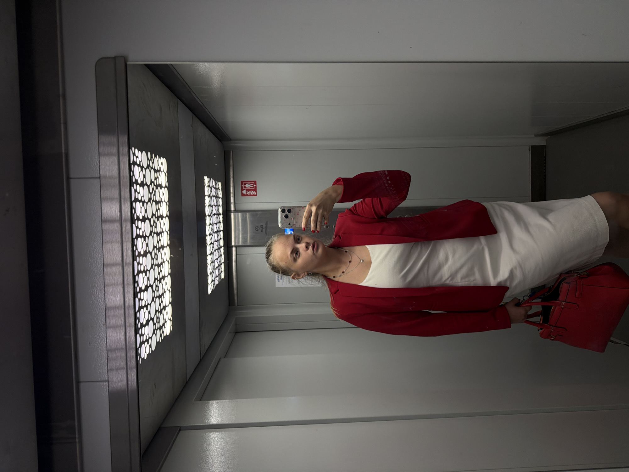 A woman is taking a selfie in an elevator. She is wearing a white dress and a red blazer, and holding a red handbag. The elevator has perforated metal panels with round cutouts. The elevator's interior is white. A sticker with the silhouette of a person and flames can be seen on the back wall of the elevator. The location appears to be Izhevsk, Russia, but that cannot be confirmed from the image.