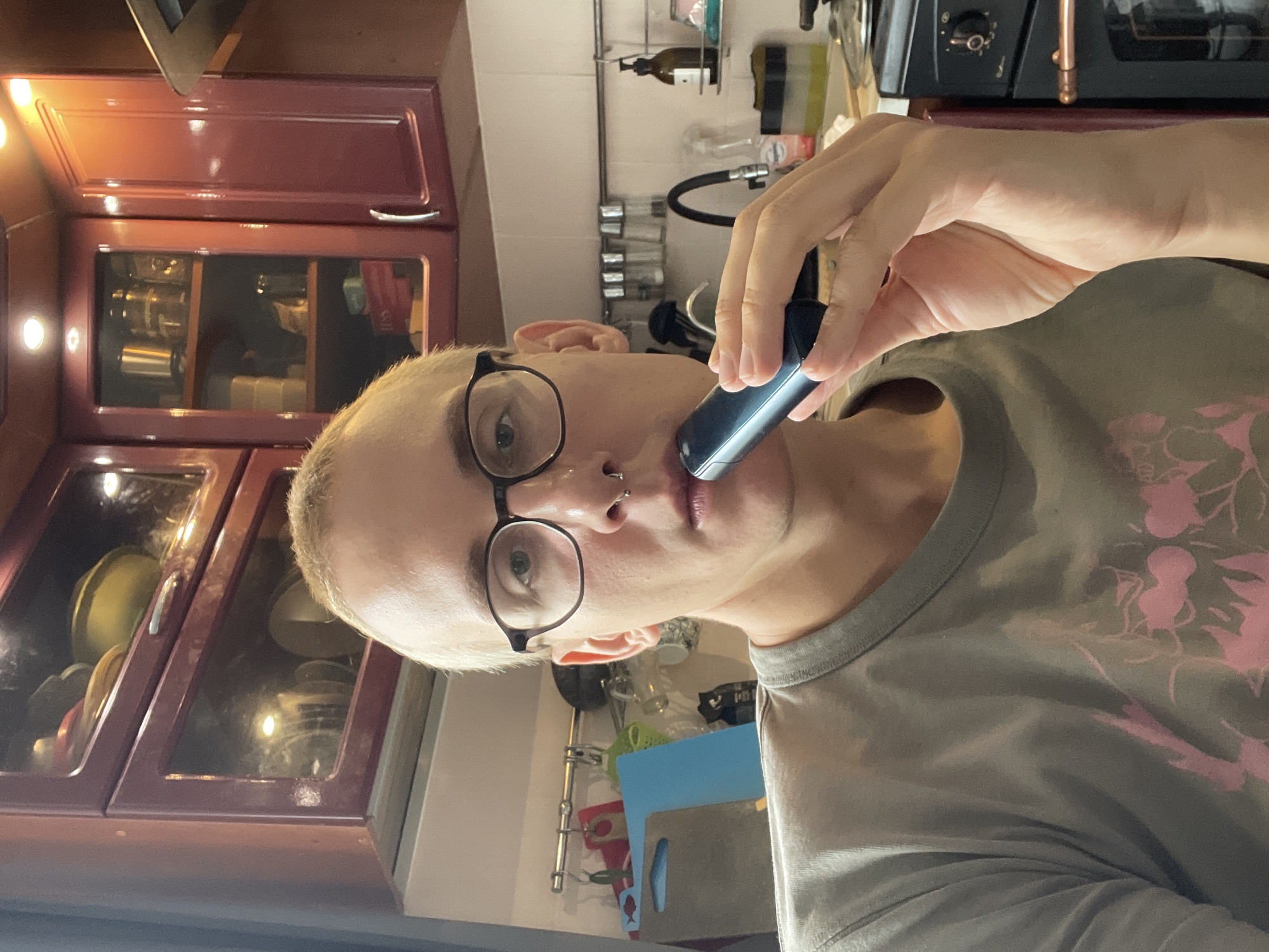 A person with short blonde hair and glasses is holding a dark blue vape pen to their lips. They are wearing a grey t-shirt with a pink floral design. The background shows a kitchen setting with maroon cabinets filled with dishes and other kitchen items. There's a sink with a faucet and various cleaning supplies. An oven is visible in the upper right corner. The image suggests an indoor location, potentially the person's home, but specific details linking it to Sestroretsk, Russia are absent.