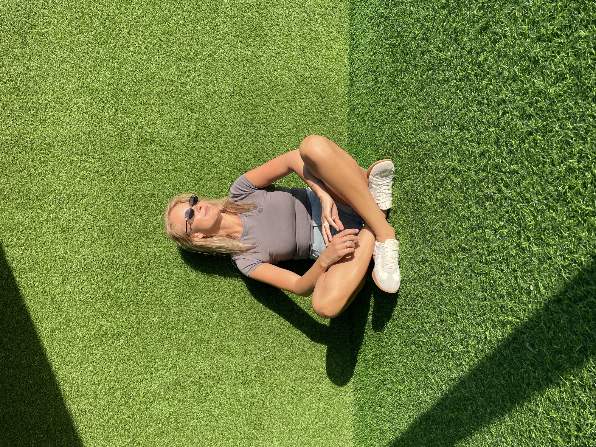 A blonde woman wearing a grey shirt, denim shorts, and white sneakers is sitting on artificial turf. The woman is wearing sunglasses. The turf is in two sections, and the shadows suggest a building corner. The image does not confirm the location is Sochi, Russia.