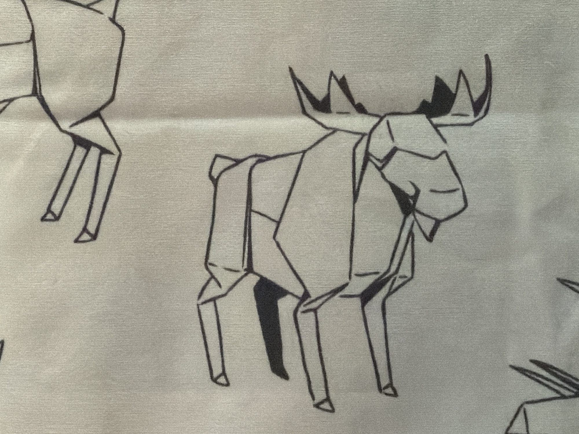 The image displays a close-up, overhead view of a fabric with a repeating pattern of black line drawings of origami animals, likely cranes, on a light grey, textured background. The origami animals are depicted in various orientations. One prominent origami animal is in the center of the frame, appearing to be in motion with its legs extended. Another partially visible origami animal is in the upper right quadrant of the image. In the lower left quadrant, there are a few vertical black lines which might represent parts of another origami animal or a decorative element. The fabric appears to be a synthetic material, possibly polyester or a blend, with a subtle weave visible. There are no people, actions, or specific environmental details visible in this cropped image.