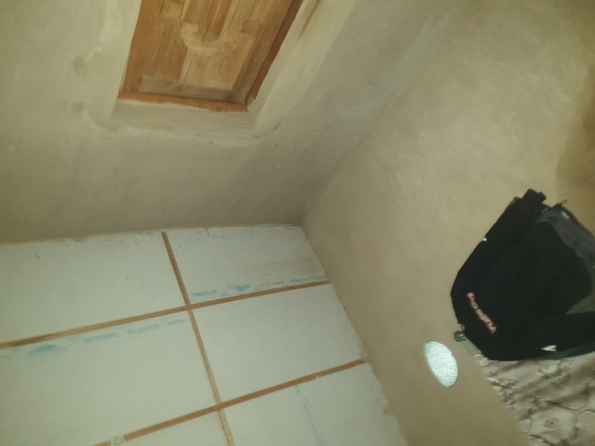 An interior corner of an unfinished room shows a plastered wall and ceiling. A wooden vent is set into the ceiling. A black sweatshirt with white and red lettering hangs from a hook on the wall. The room appears to be under construction.