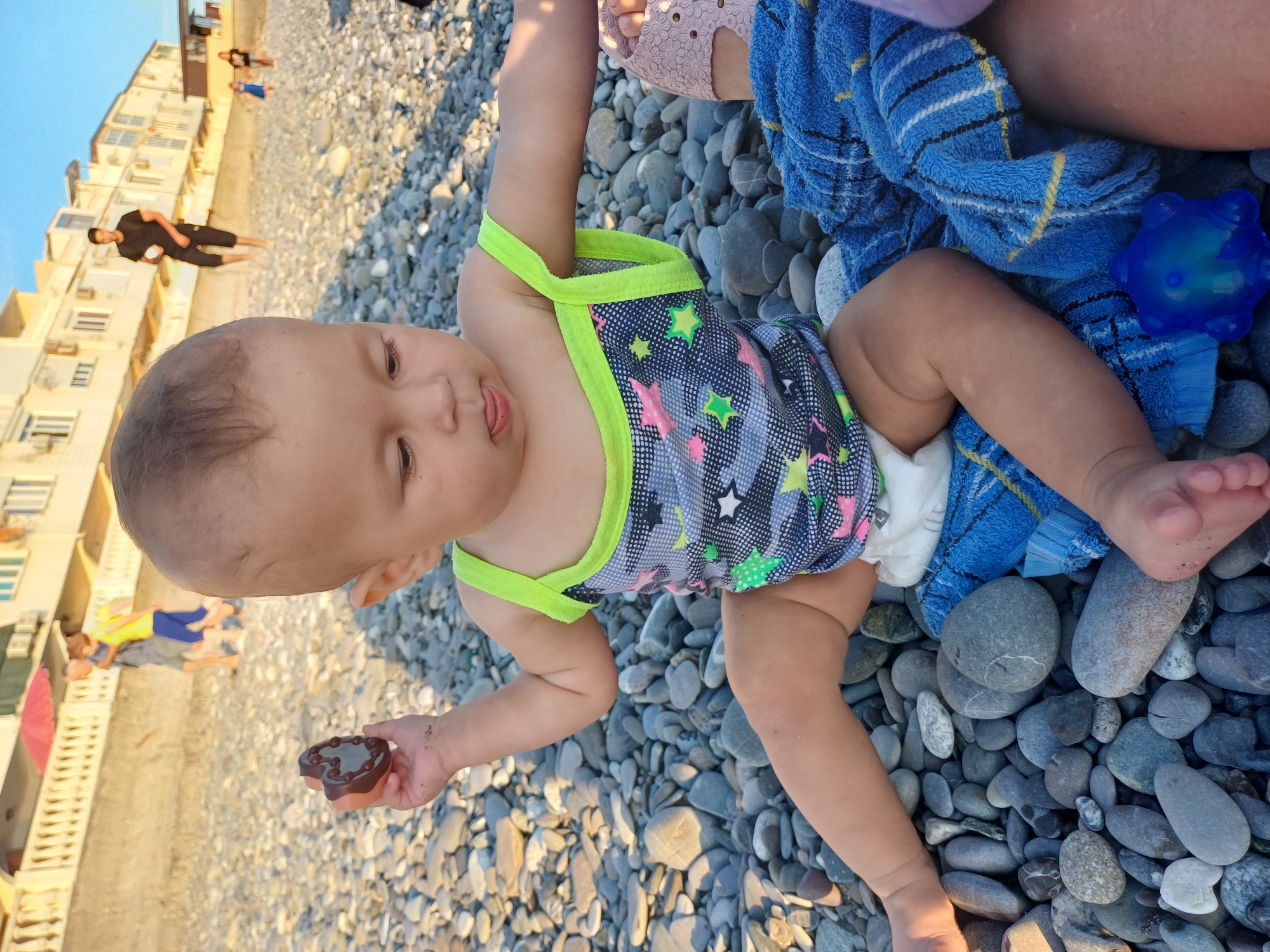 The image depicts a baby sitting on a pebble beach in Sochi, Russia. The baby is wearing a patterned swimsuit and a diaper, and is holding a chocolate-colored toy in its left hand. The baby is looking down and to the right, with its tongue slightly out. In the background, several people are walking along the beach. The buildings in the background suggest a coastal resort area. The overall scene is brightly lit, indicating it is daytime.