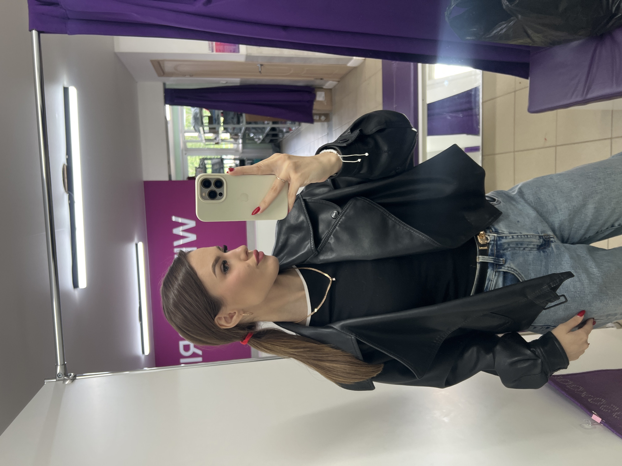 A young woman is captured in a mirror selfie, standing in what appears to be a fitting room or a similar retail space. She is seen from the waist up, holding a light-colored smartphone with multiple cameras in her right hand to take the photo. Her hair is styled in a low ponytail, secured with a red elastic band, and she wears a black leather jacket over a black top, paired with blue jeans. A gold necklace and gold earrings are visible, along with a ring on her right hand and red nail polish. Her expression is a subtle pout, as she looks towards the phone.
The setting is an indoor area with white walls and a large mirror that reflects the woman and parts of the room. Purple curtains are visible at the top and side of the mirror, suggesting changing room dividers or decor. In the reflection of the mirror, beyond the woman, a window is visible showing green trees and natural light, indicating it is daytime. Strip lighting fixtures are mounted on the wall above the mirror. Part of a purple sign or banner with white, mirrored, and upside-down text "W" and "BI" can be seen on the wall behind her in the reflection. The overall impression is a casual pose within a commercial environment.