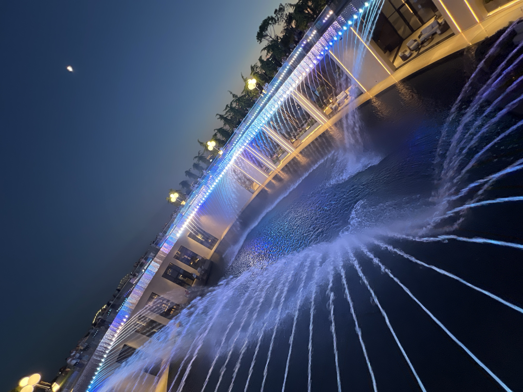 The image captures a view of the "Chistie Kluchy" (Pure Springs) fountain complex in Krasnodar, Russia, at dusk. The central feature is a multi-tiered fountain with numerous streams of water cascading downwards, illuminated by blue and purple LED lights. The fountain is situated around a contemporary building with large windows and outdoor seating areas. A walkway with decorative lighting runs along the upper level of the fountain. In the background, trees and streetlights are visible against a deep blue sky, with a crescent moon faintly visible. Several people are present on the walkway, observing the fountain. The perspective is from above and to the side, emphasizing the dynamic flow of water and the architectural design of the complex.