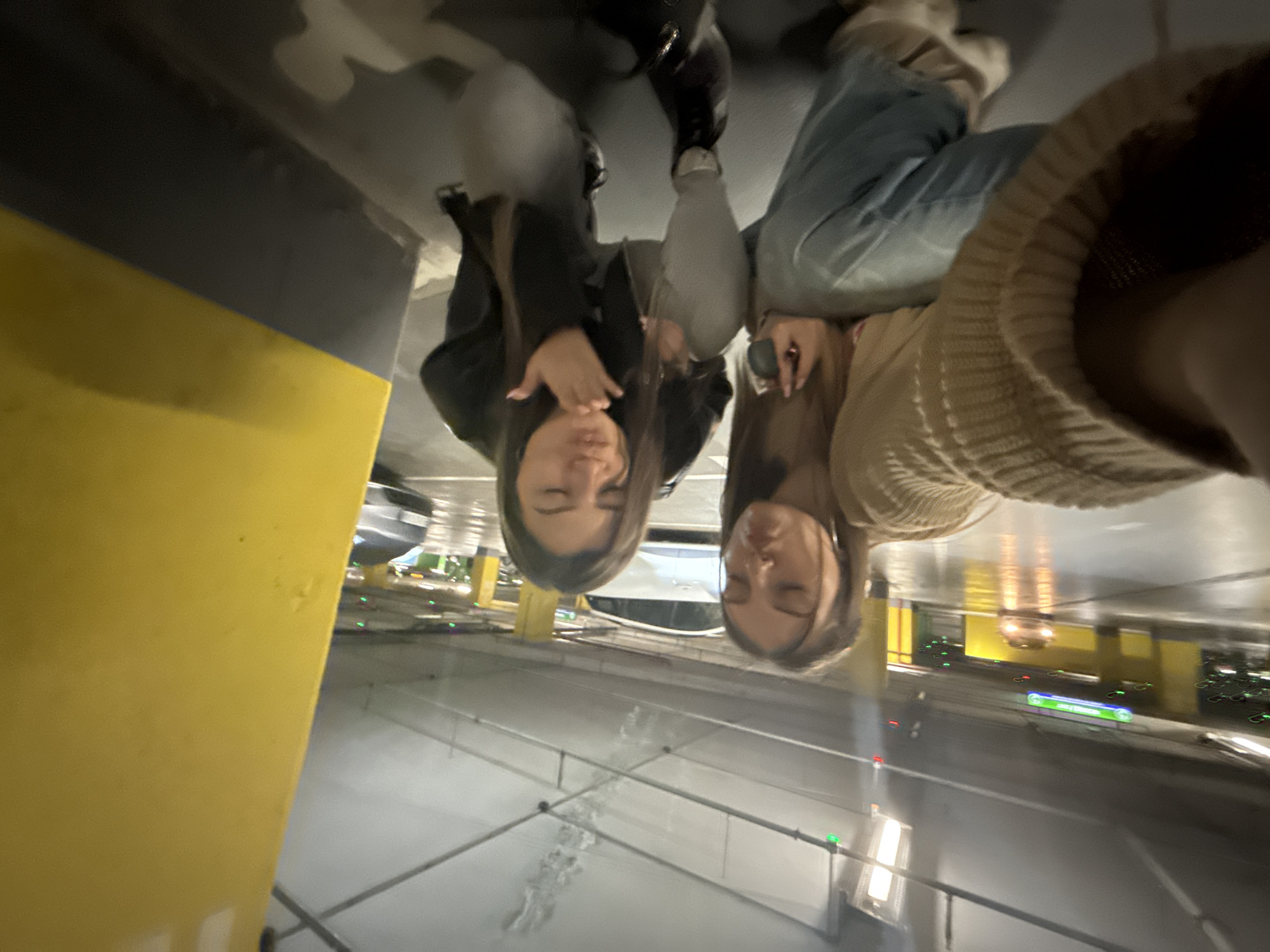 Two women are reflected in a wet floor in an underground parking garage. The woman on the left wears a dark jacket and black boots. The woman on the right wears a light-colored sweater and blue jeans with beige socks. The parking garage has yellow support columns and overhead lighting. A sign reading "AREA 51 Parking" is visible. The city is Sofia, Bulgaria.