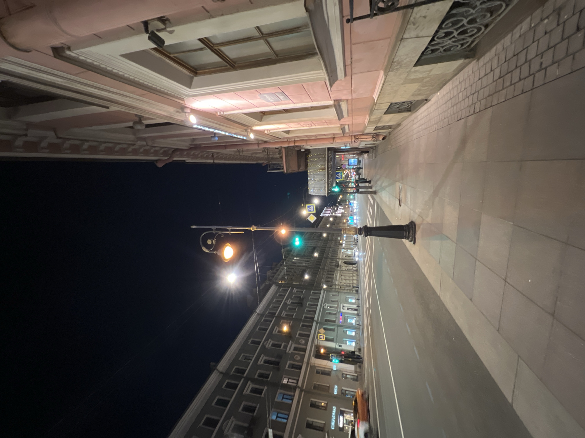 The image captures a nighttime street scene in an urban setting, likely Saint Petersburg, Russia. The perspective is looking down a city street, with buildings on both sides, a sidewalk, and a road.
On the left, a multi-story building facade, possibly light pink or pale orange, displays classical architectural details including cornices, decorative moldings, and recessed areas illuminated by spotlights. The building appears well-maintained, with some windows visible above.
In the middle ground, a traditional black lamppost with ornate scrollwork holds two lamps emitting a warm, golden-orange light. Further down the street, additional streetlights with cooler white light and a green traffic light contribute to the illumination.
The street itself consists of a wide asphalt road with white lane markings, indicating traffic lanes. Adjacent to the road is a paved sidewalk, featuring large grey square tiles and a section with a smaller, brick-like pattern.
Across the street, more multi-story buildings are visible, characterized by numerous windows, some of which are lit, creating a distant cityscape. The sky is completely dark, suggesting a clear night.
In the distance, blurry figures of people can be seen on the sidewalk, indicating some pedestrian activity. Faint streaks of light in the far background suggest the movement of vehicles. While some street signs are visible (e.g., a yellow diamond shape), specific text on buildings or signs is largely unreadable due to distance and blur. The scene conveys the quiet ambiance of a city street at night, brightly lit by artificial sources.