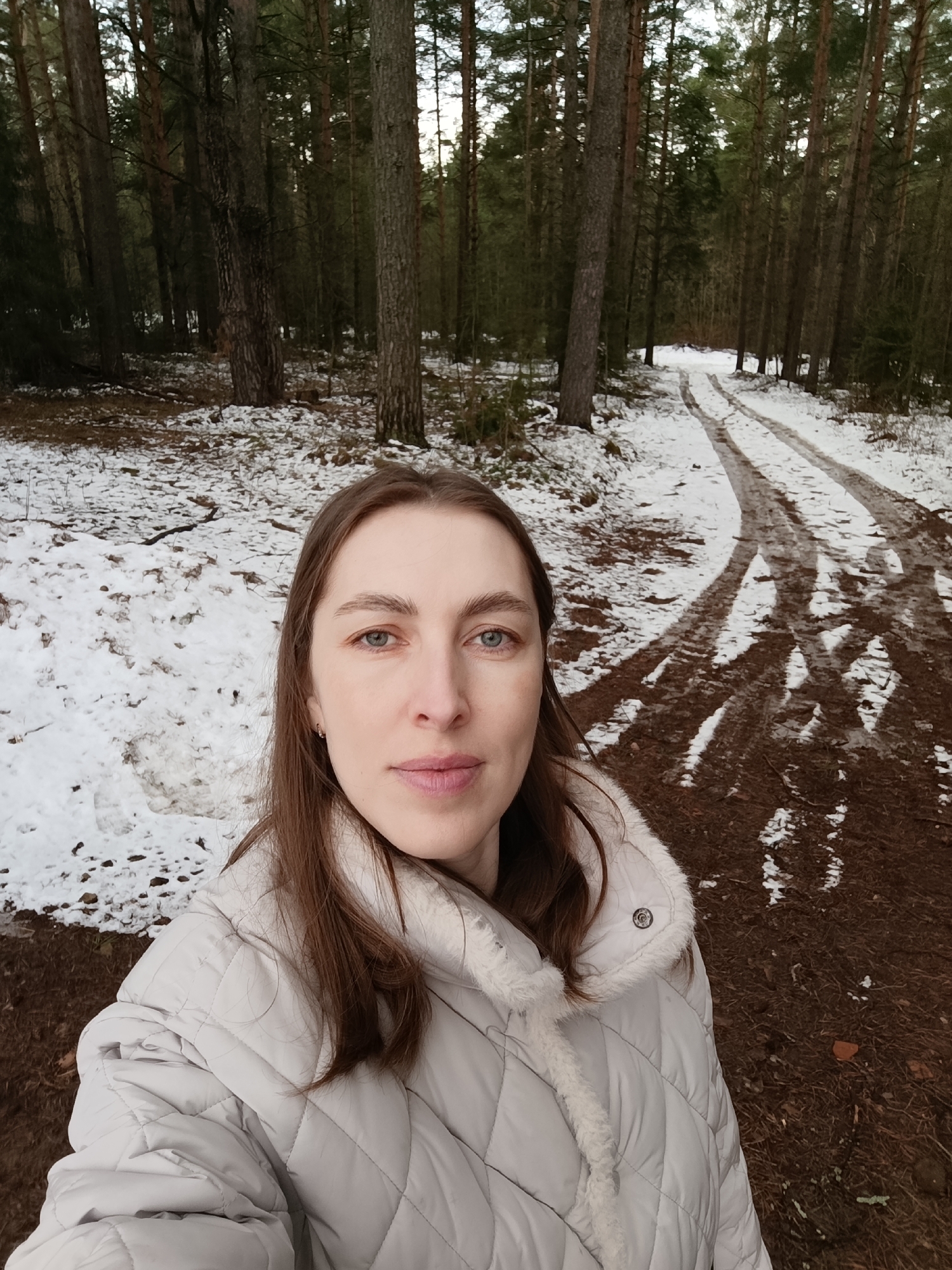 A woman is taking a selfie in a snow-covered forest in what is likely the city of Chaikovsky, Russia. She has light skin, brown hair, and blue eyes, and is wearing a light-colored quilted jacket with a fur-trimmed collar. Behind her is a dirt road with tire tracks leading through the woods. Patches of snow are visible on the ground among the trees. The trees are tall and bare, suggesting it is winter or early spring.