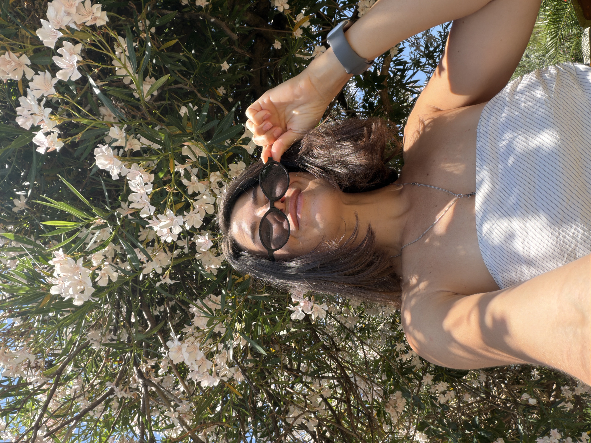 The image shows a woman lying on her back in a garden, with her head propped up on her arm. She is wearing a white, pinstriped top and sunglasses. Her hair is dark brown and appears to be styled in loose waves. A silver necklace is visible around her neck. She is holding a pair of dark-rimmed sunglasses in her hand, as if about to put them on. The background is filled with lush green foliage and white flowers, suggesting a sunny day and a warm climate. The sunlight casts dappled shadows on her skin and clothing, indicating it is daytime. The overall mood is relaxed and casual, with the woman appearing to be enjoying the outdoor scenery. The orientation of the image is vertical, with the woman's head towards the bottom and her feet towards the top.