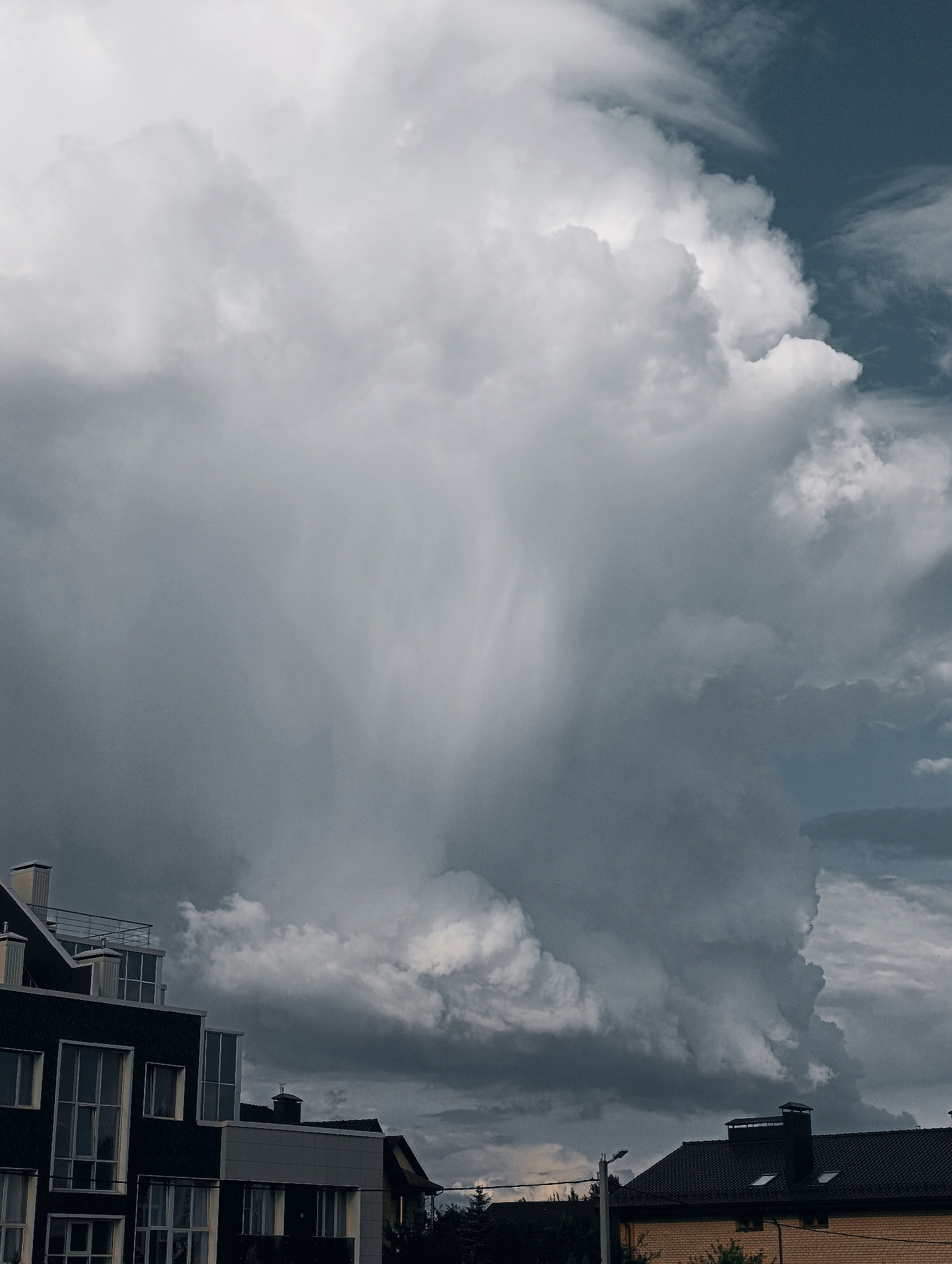 A photograph captures a dramatic sky filled with a towering cumulonimbus cloud formation. The cloud is predominantly white and gray, with darker, denser sections suggesting an impending storm. Streaks of rain or virga are visible descending from the cloud mass. In the foreground, the rooftops of several buildings are visible. The building on the left is dark-colored with numerous windows, some of which are illuminated. The building on the right is lighter in color with a dark tiled roof and chimneys. The overall scene suggests a threatening weather event approaching a residential or urban area. The location is Unknown, Unknown.