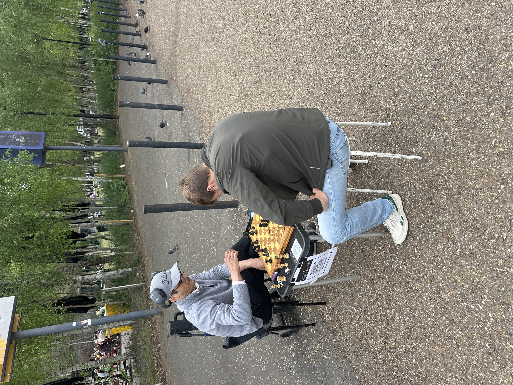 Two men are playing chess outdoors. One man sits on a rolling chair, wearing a grey Titleist hat, glasses, headphones, a grey sweater, and black pants. The other man is standing and leaning over the chessboard, wearing a dark green jacket and blue jeans. The chessboard is placed on a small platform between the two men. The location appears to be in a park in London, United Kingdom, as evidenced by the row of black bollards separating a paved walkway from a grassy area with trees, and a sign. There are pigeons in the background.
