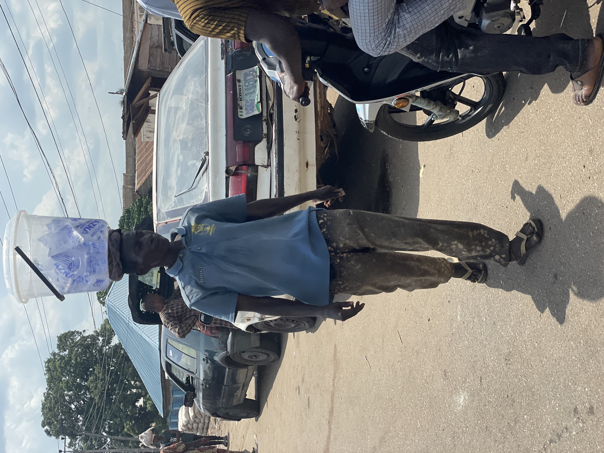 In Lokoja, Nigeria, a man in a blue shirt and stained brown pants walks on a paved road carrying a bucket full of water sachets on his head. He wears sandals. Various vehicles surround him, including a white van, a dark blue car, and a motorcycle. Power lines are visible above. Other people are also in the vicinity. The license plate on the white van reads "KOGI KRE-114BG."