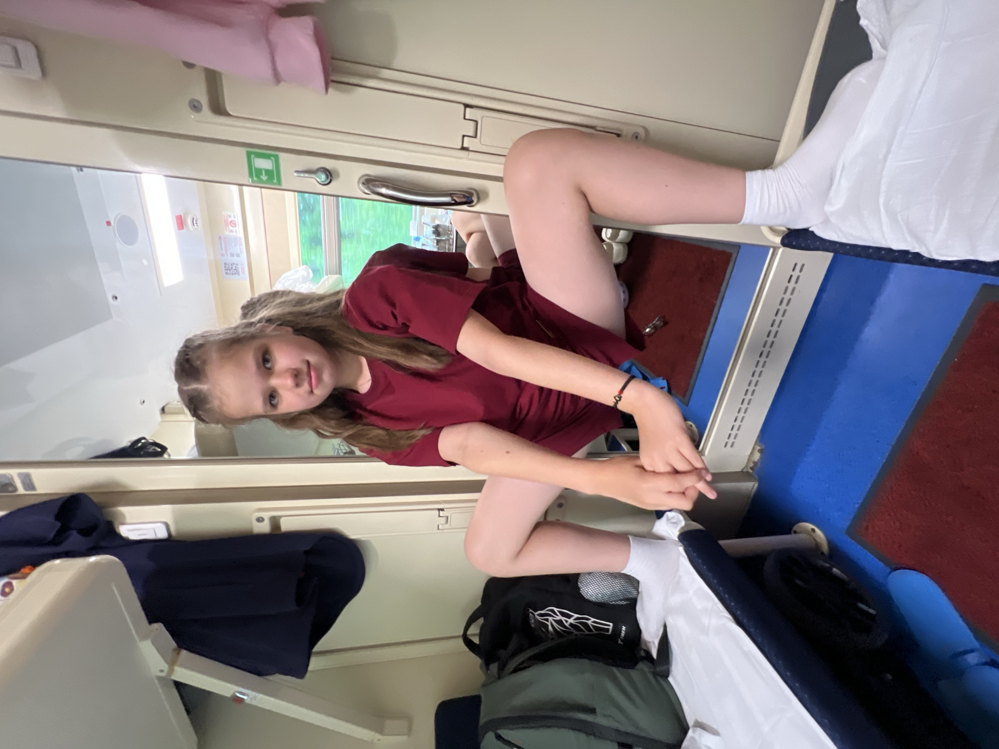 A young girl with long blonde hair, styled in braids, is positioned in a seated, kneeling pose within a train compartment. She is wearing a dark red t-shirt and white socks. Her expression is direct and engaging, looking towards the camera. Her arms are crossed and resting on her knees. The compartment features typical train interior elements such as overhead luggage racks, a window showing greenery outside, and bunk beds. One bunk is covered with white bedding, suggesting it is occupied or prepared for sleep. Dark blue fabric is visible on a lower bunk or seat, and several bags, including a green backpack, are stowed in the compartment. The lighting suggests it is daytime. The overall atmosphere is casual and suggests a journey in progress.