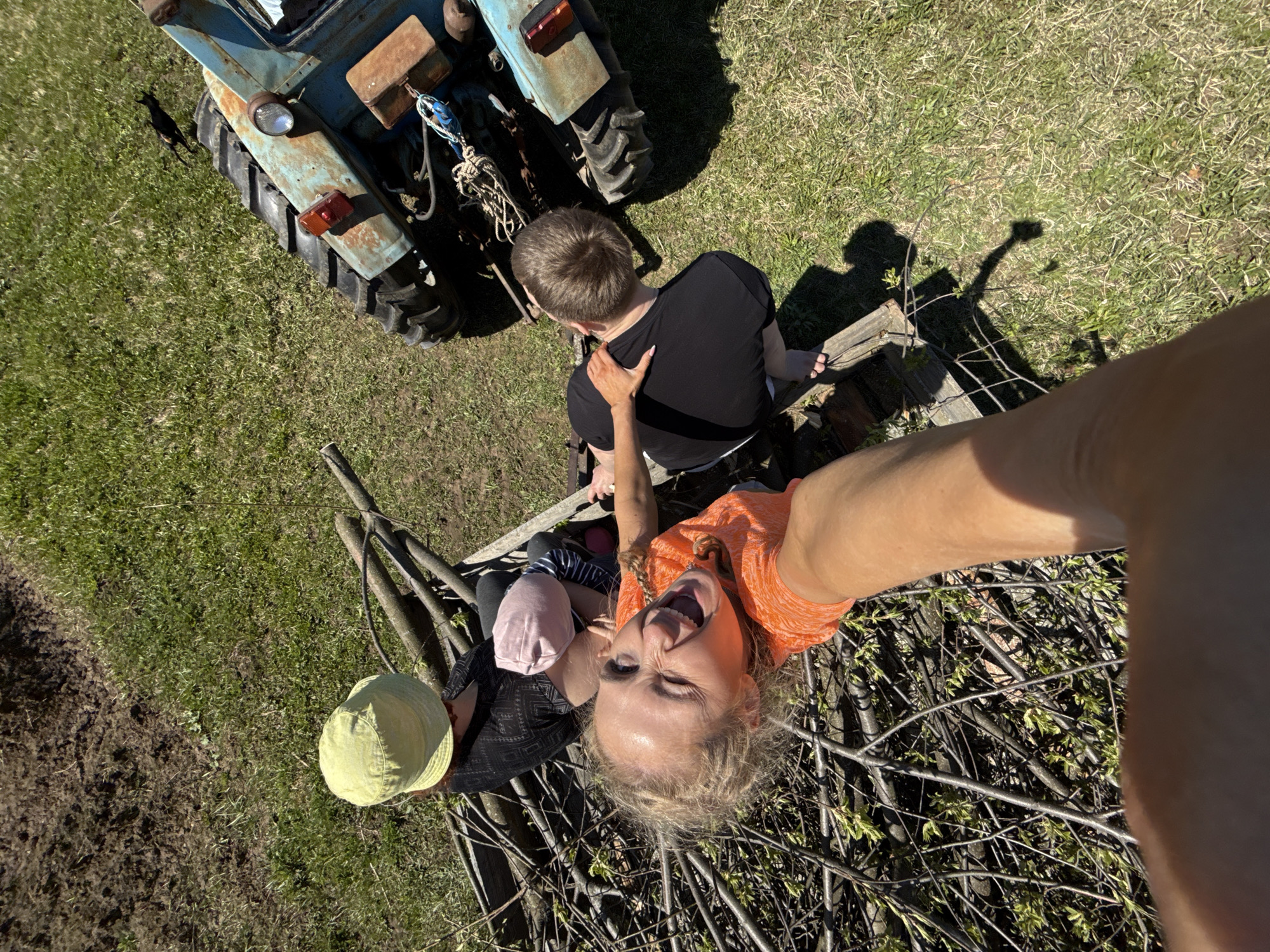 The image shows three people and an old blue tractor in an outdoor setting in what appears to be a grassy area. A person in an orange shirt is taking a selfie, with the camera angled downward. To the left is a person with a light green hat, and a toddler in a pink hat. There is a man wearing a black shirt sitting down on a wooden fence near the tractor, with the hand of a person (presumably the person taking the selfie) touching his back. The environment is primarily grass with some dirt patches visible, with some sticks and branches scattered near the lower portion of the image. The location is Nizhnyaya Chermoda, Russia.