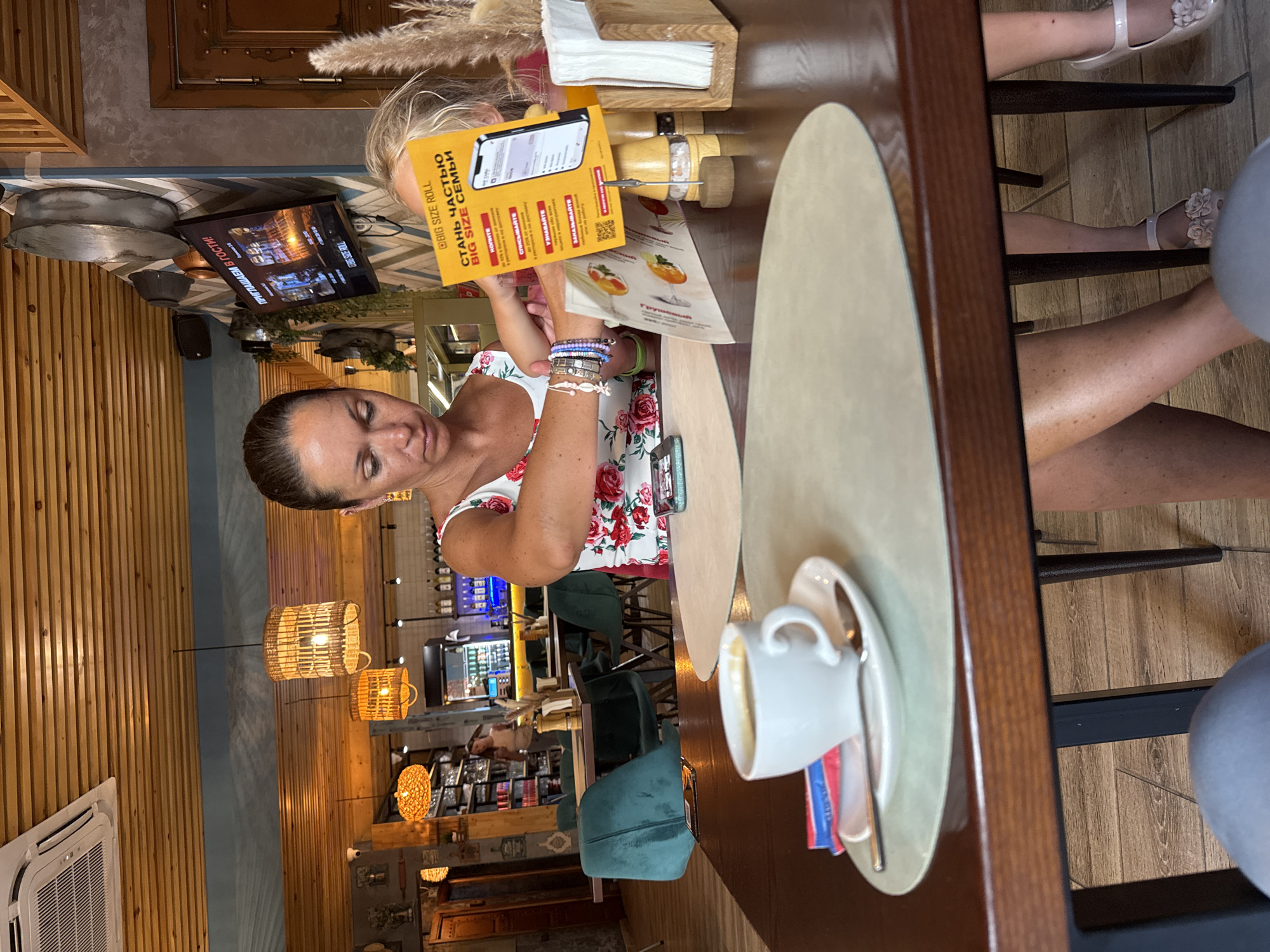 In Sochi, Russia, at a restaurant, a woman with dark hair pulled back is seated at a table. She is wearing a white dress with a floral pattern and is looking down at a menu she is holding. She has several bracelets on her left wrist. In front of her is a white cup and saucer with a spoon, as well as two packets of sugar or sweetener. The table is dark wood, and a light-colored placemat is on it. Behind her, a blonde child's head is visible. In the background, the restaurant interior is visible, with wooden paneling on the ceiling and walls, woven light fixtures, shelves stocked with items, and other tables and chairs. A TV screen is mounted on the wall, displaying an advertisement.