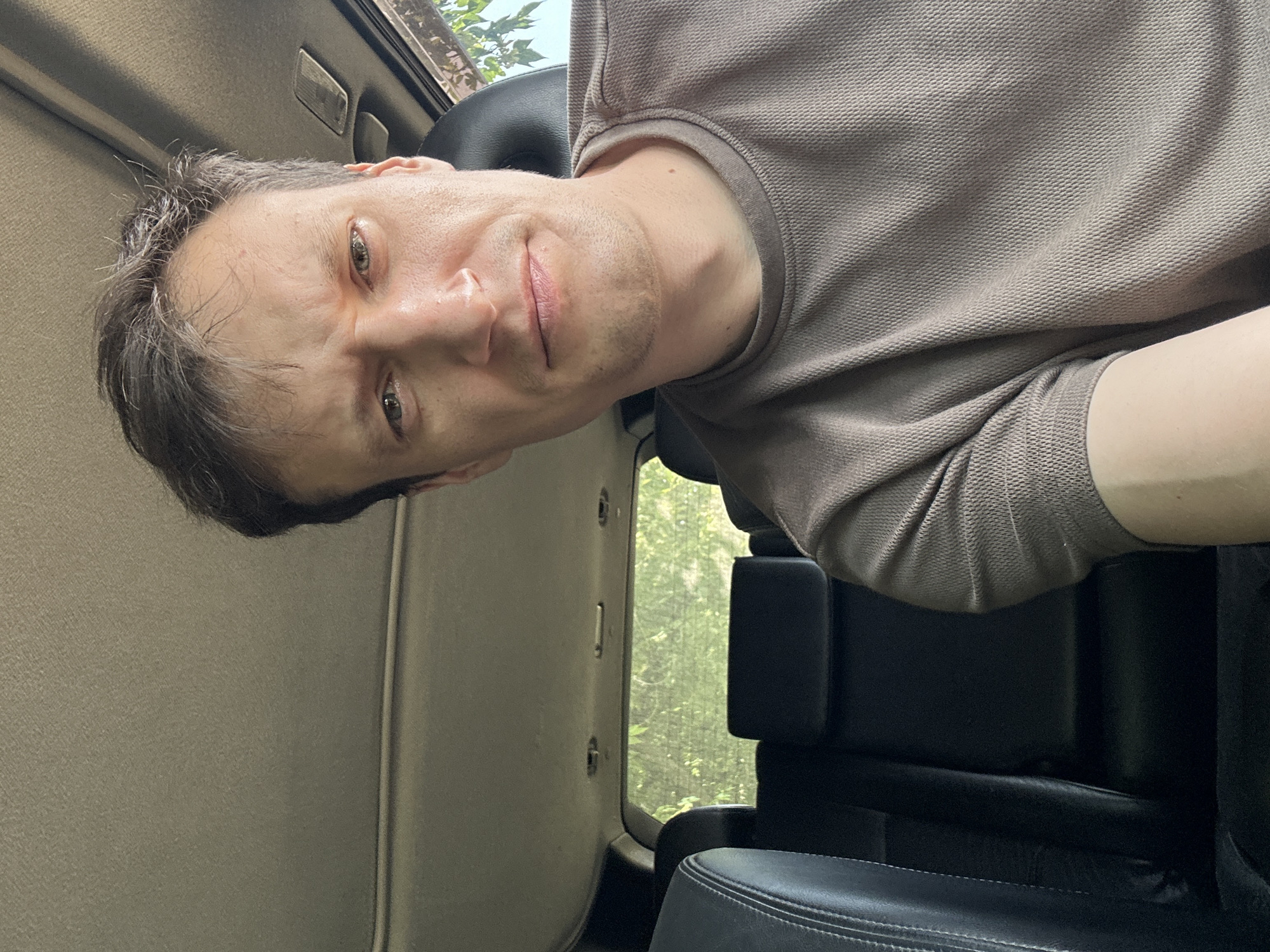 A close-up, eye-level, selfie-style shot shows a man with short, dark hair and light skin sitting in the front passenger seat of a car. He is looking directly at the camera with a slight frown. The man is wearing a brown, textured t-shirt. The background shows the interior of the car, including the dark leather seats in the back and the car's headliner. Through the rear window, trees and foliage are visible, suggesting an outdoor environment, potentially a rural or suburban area, but no specific city or location can be identified from the image itself.