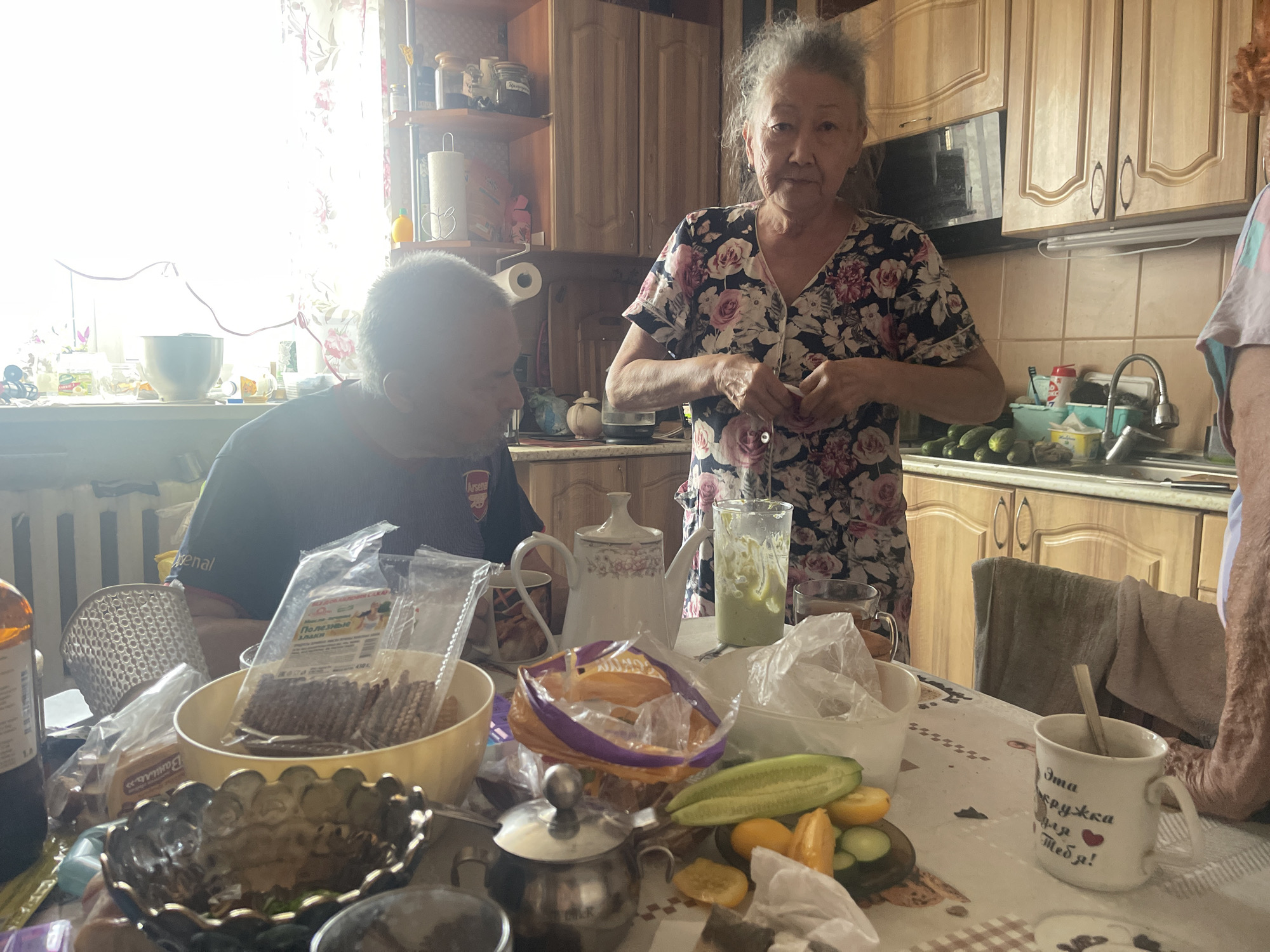 The image depicts an indoor scene, likely a kitchen or dining area, in the city of Sibirskiy, Russia. The lighting suggests it is daytime, with bright light coming from a window on the left side of the frame, partially obscuring the background. In the foreground, a table is laden with food and kitchenware. Visible items include a yellow bowl containing packaged snacks, a decorative metal bowl, various jars and containers, a white teapot with floral patterns, a glass with a green liquid, and cut vegetables such as cucumbers and tomatoes. A white mug with Cyrillic text and a heart symbol is also present, with a spoon inside.
Two individuals are partially visible. On the left, a man with graying hair is seated, wearing a dark t-shirt with a red emblem on the shoulder. His gaze is directed downwards, and he appears to be holding something in his hands. On the right, an older woman with gray hair is standing, wearing a floral-patterned dress. She is looking towards the camera and holding a small red item in her hands. Her expression is neutral.
The background features kitchen cabinets with wooden doors and tiled backsplash. Shelving units containing various jars and containers are also visible. A radiator is present on the far left wall. The overall impression is a casual domestic setting, possibly during a meal. The presence of Russian text on the mug and packaging confirms the location as Russia.