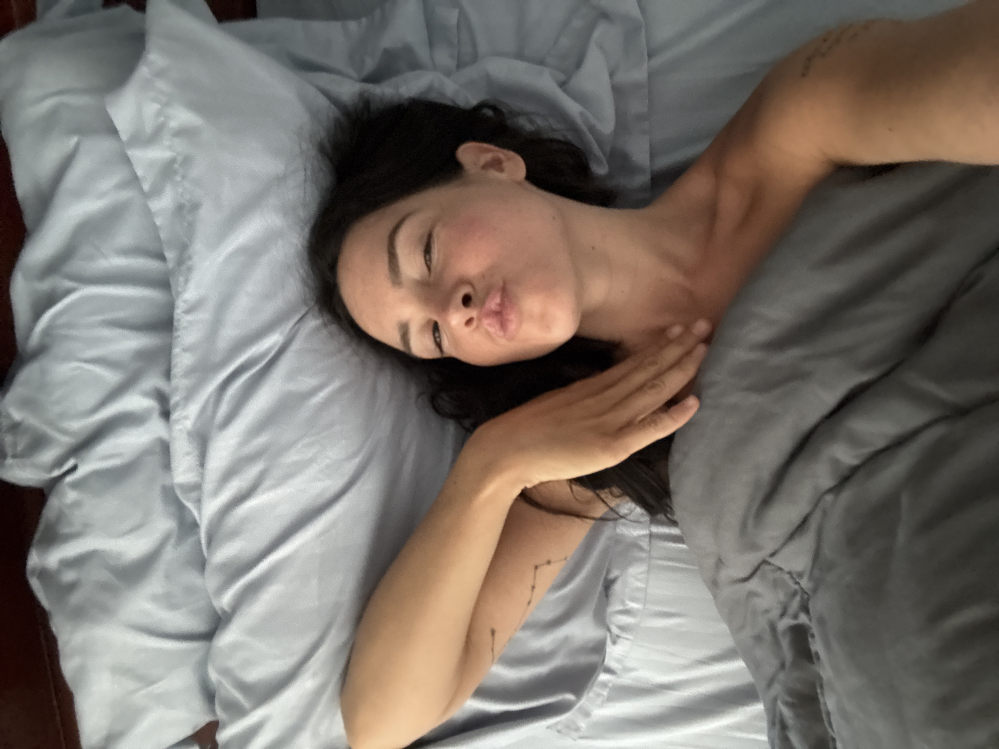 An overhead, eye-level, medium shot captures a woman with dark hair lying on a bed with pale blue sheets in North Kuta, Indonesia. She is facing the camera, with her head tilted to her right, and her lips pursed in a kiss. Her right arm is bent, with her hand resting on her neck and chest, partially covered by a dark gray comforter. A small, linear tattoo is visible on her upper left arm. The lighting suggests a soft, ambient light source, likely from a window, creating subtle shadows on the fabric and her skin.
