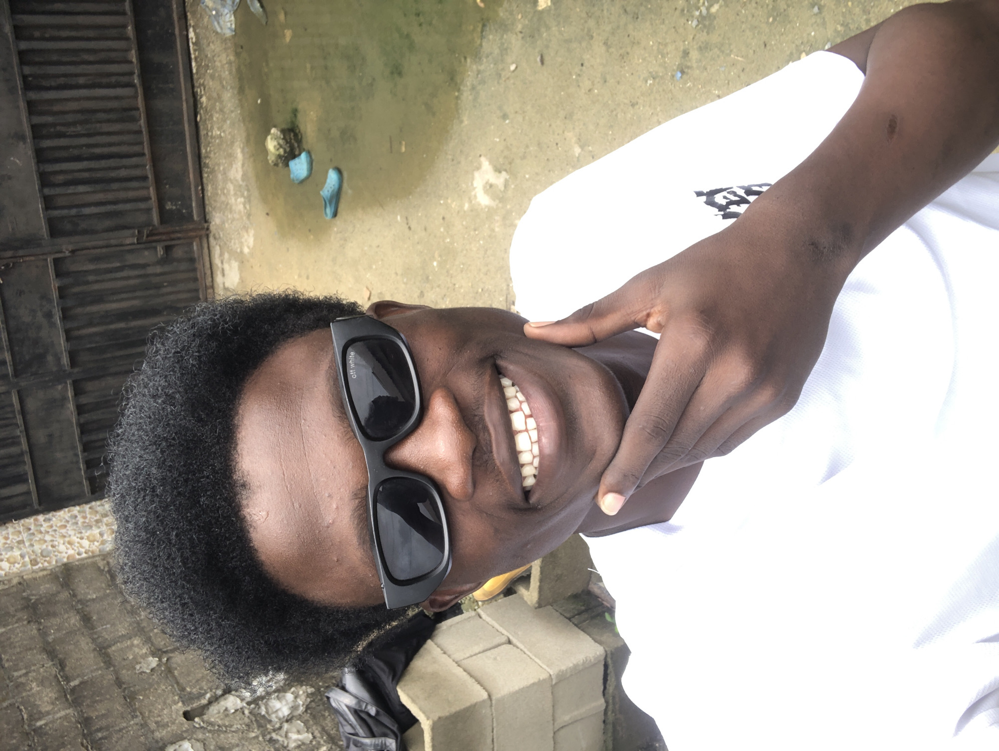 This is a medium-shot, eye-level, outdoor selfie of a young Black man in Ado, Nigeria. The man has dark skin, a short Afro hairstyle, and is wearing black sunglasses and a white t-shirt. He is smiling broadly and has his right hand on his chin. Behind him, there is a textured concrete wall, a dark metal gate, and a muddy, wet ground with two blue flip-flops. The man's posture and smile suggest a casual and happy demeanor. The overall setting appears to be an urban or semi-urban environment.