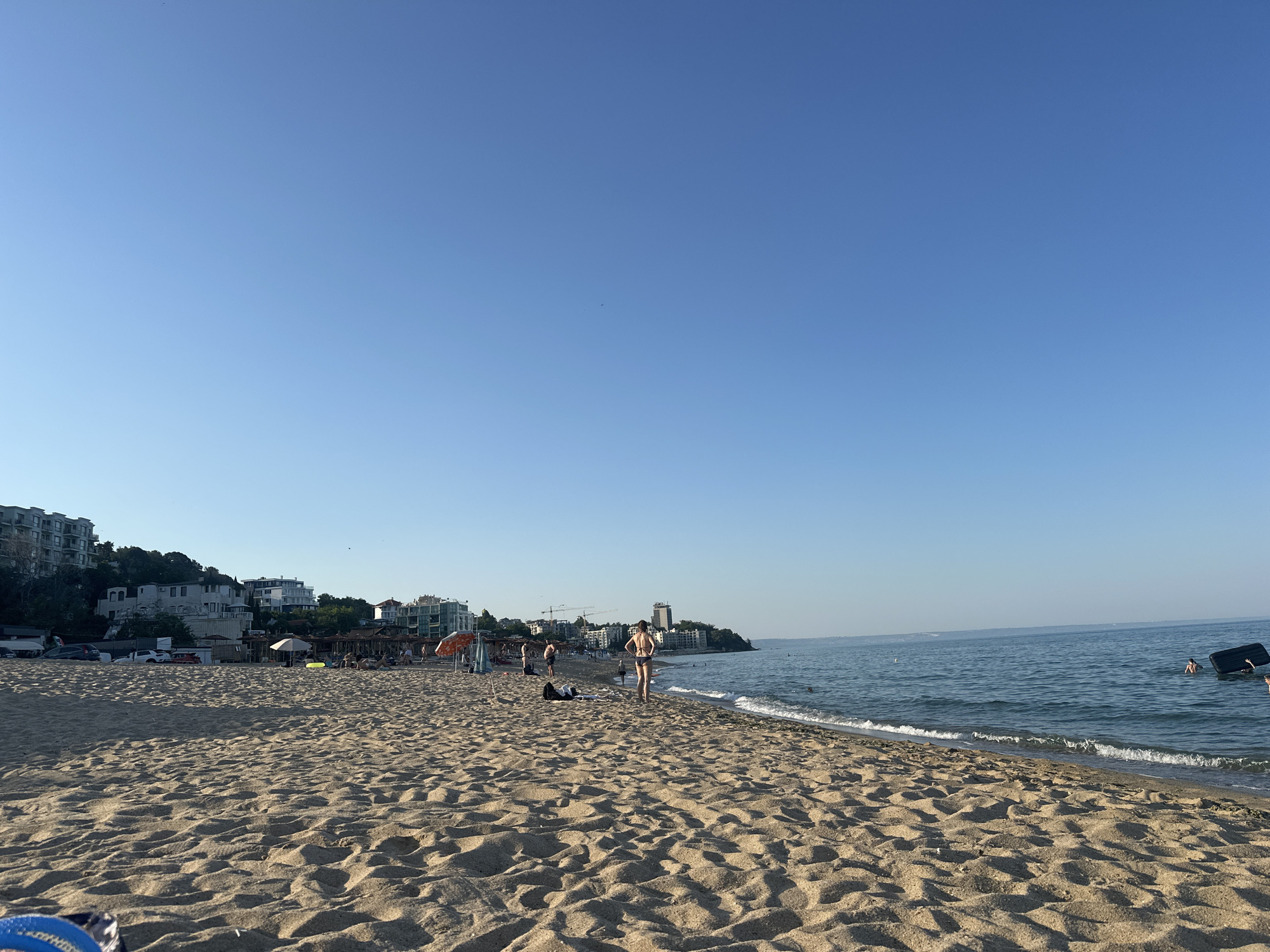 The image depicts a sunny day at a sandy beach with a clear blue sky. Several people are visible on the beach and in the shallow water. Some are sunbathing or relaxing under umbrellas, while others are wading or swimming. A few buildings are situated on a hill overlooking the beach, suggesting a coastal town or resort. The sand is rippled from the wind and the gentle waves are lapping at the shore. The overall atmosphere appears relaxed and leisurely, consistent with a vacation or a day at the beach. The time of day seems to be late afternoon or early evening, given the warm, soft lighting.