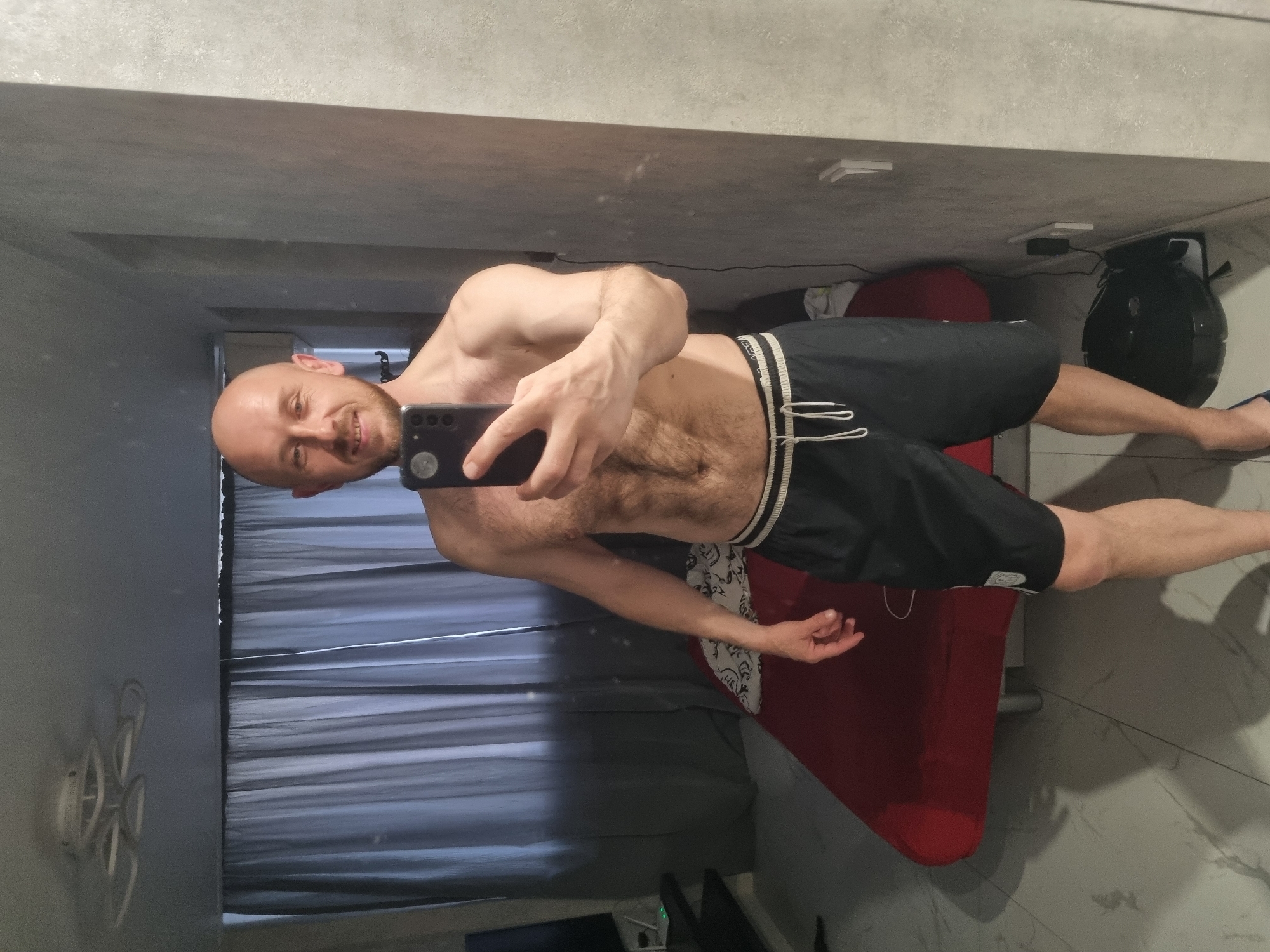 A shirtless, fair-skinned male with a shaved head is taking a selfie indoors. He is wearing black athletic shorts with white stripes along the side and white drawstrings. He's holding a black smartphone in his right hand. The background includes a curtained window, a red padded bench, a black motorcycle helmet, and tiled flooring. The location in the city of Sochi, Russia is not evident from the image.