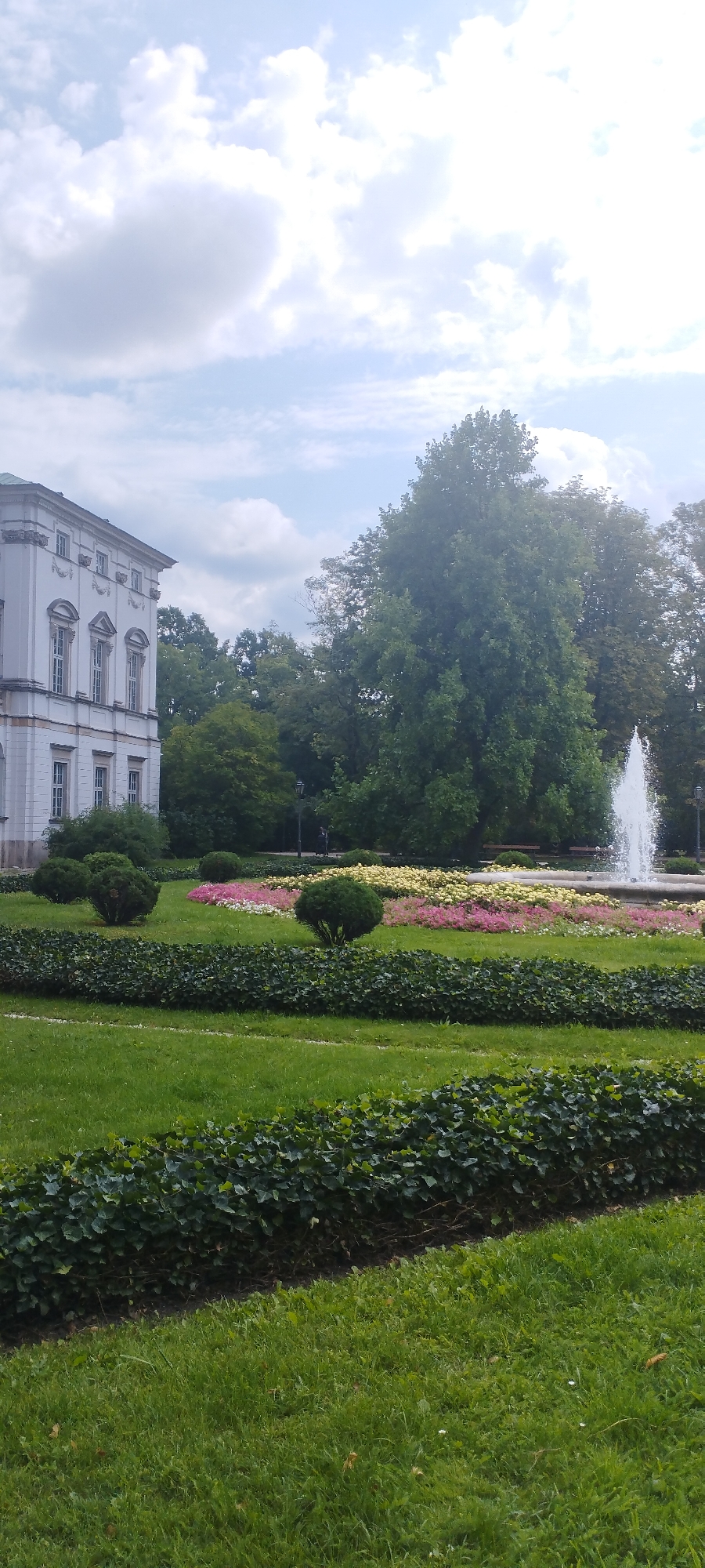 This is a vertical, eye-level, medium shot of a formal garden in front of a white, classical building. The building, which is on the left side of the frame, has multiple windows and ornate architectural details. In the center of the image, a vibrant flower bed with pink, white, and yellow flowers is visible. Beyond the flower bed, a tall fountain sprays water into the air. Lush green trees and hedges form the backdrop of the scene. The sky is partly cloudy with patches of blue. The scene appears to be a peaceful and well-maintained public space. The style of architecture suggests a historical or governmental building, possibly a palace or museum, located in Warsaw, Poland, which is known for its beautiful gardens and classical architecture. The image captures a serene moment, with no people clearly visible in the foreground.