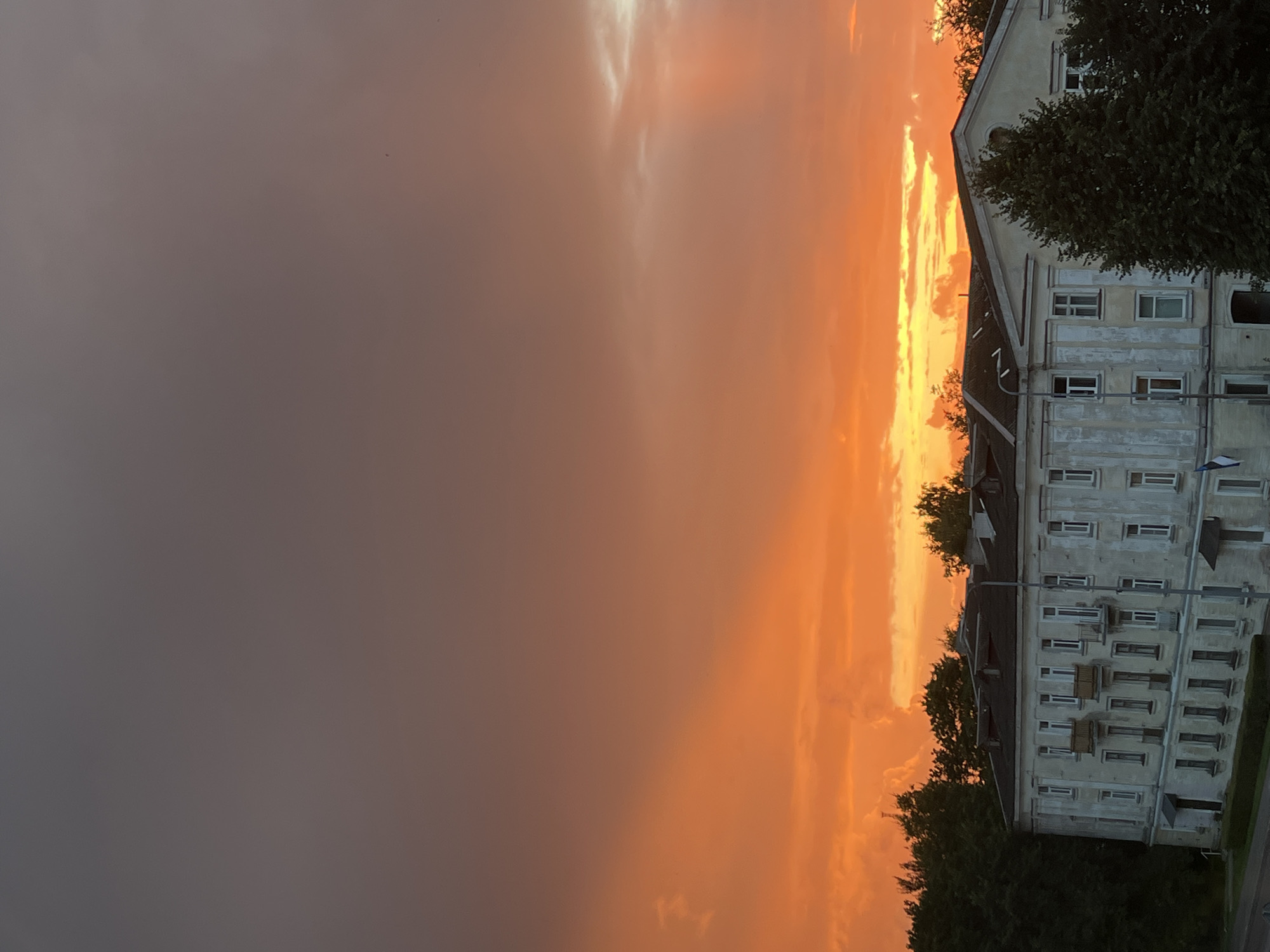 The image depicts a dramatic sunset over a Soviet-era apartment building in Kohtla-Järve, Estonia. The sky is a vibrant gradient of orange, yellow, and deep gray, with textured clouds catching the last rays of sunlight. The apartment building, with its pale, weathered facade and symmetrical rows of windows, occupies the lower half of the frame. Dark green trees flank the building and a lamppost stands in front of it. A small Estonian flag is visible on the left side of the building. The scene is devoid of people and any observable action.