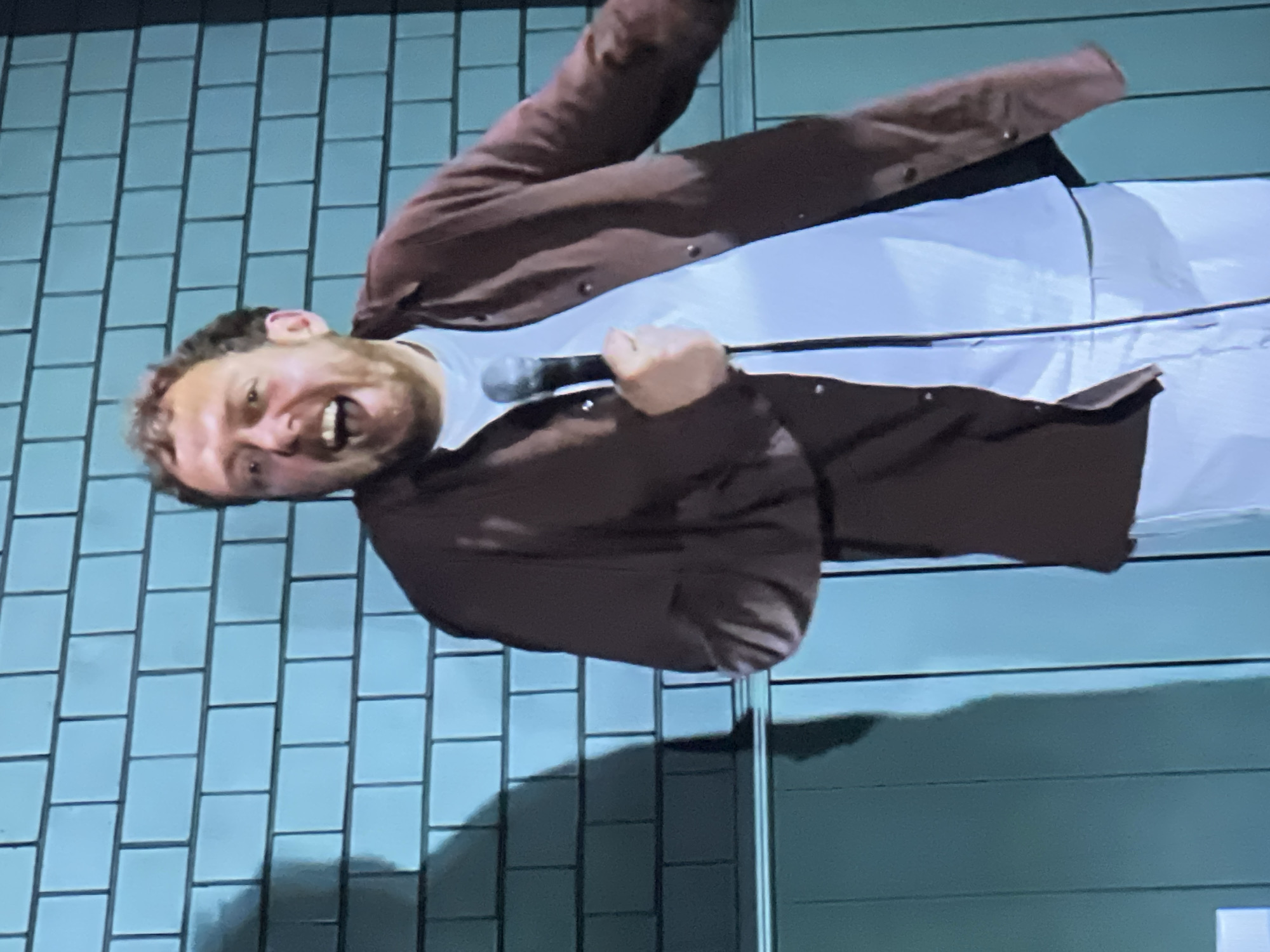 The image shows a man with curly brown hair and a beard standing in front of a light blue tiled wall, holding a microphone. He is wearing an open, dark brown shirt over a white t-shirt and white pants. He is looking towards the camera with his mouth open and appears to be speaking or singing. A shadow of the man is cast on the wall to his left. The lower part of the image shows a vertical teal surface. There is no identifiable information in the image to suggest it is located in Đà Nẵng, Vietnam.