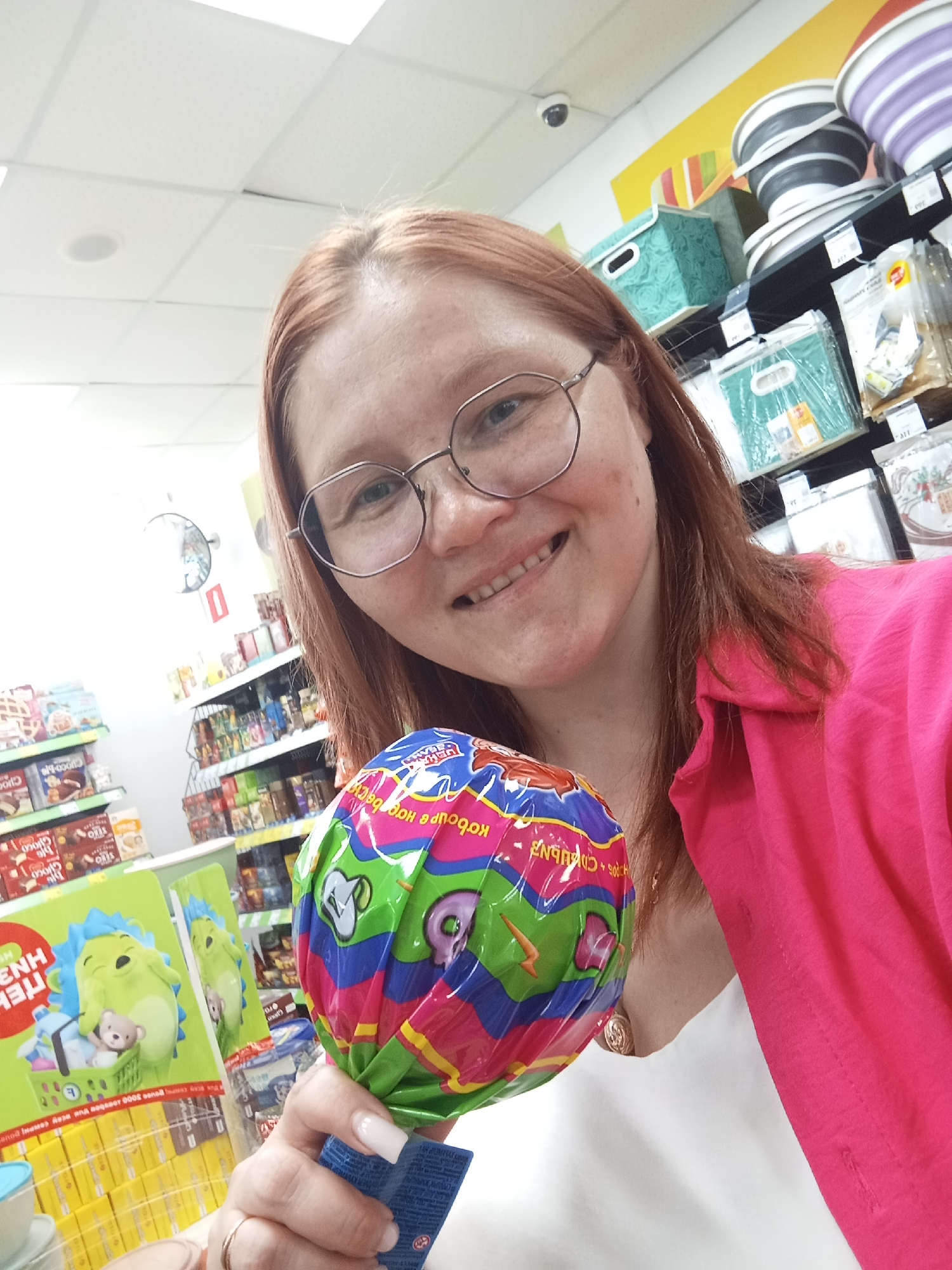A woman with reddish hair and glasses is holding a large, wrapped lollipop. She's wearing a pink shirt over a white top and has a gold ring on her left ring finger. She is smiling and holding the lollipop towards the camera. The background appears to be a store aisle with shelves stocked with various goods. In the bottom left corner, there is a green cardboard display with a cartoon hedgehog character. Above her are shelves with storage containers and other items. A security camera is visible on the ceiling. The exact location is unknown, but it is somewhere in the city of Unknown, Unknown.