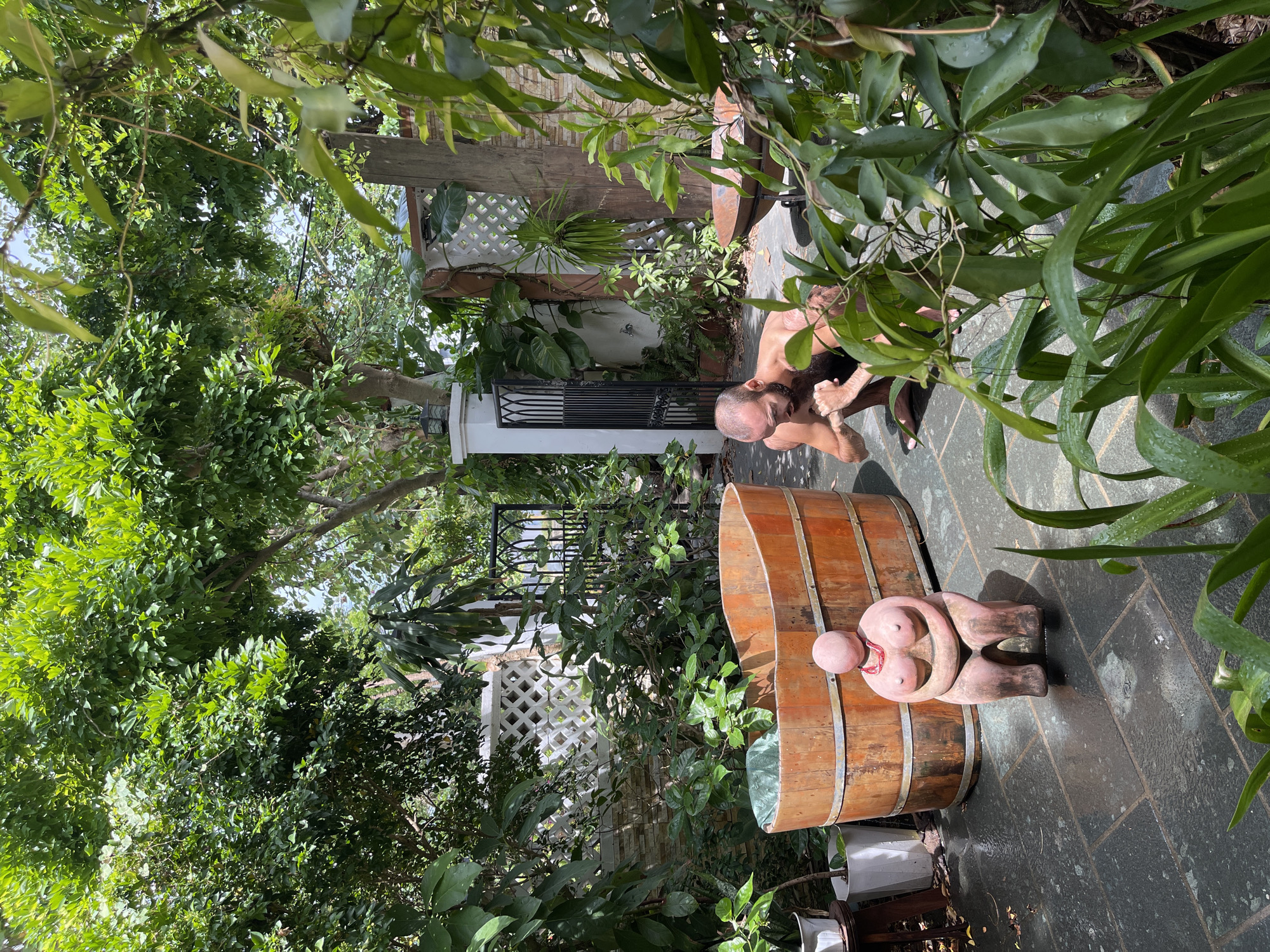 In the city of Đà Nẵng, Vietnam, a naked man with a beard is crouching down in a lush green garden. He appears to be tending to something on the ground. To his left is a large wooden tub and a pink, stylized sculpture of a woman. The garden features stone paving, various plants, trees, and white lattice fencing. A black metal gate is visible in the background. The scene suggests a private outdoor space, possibly a backyard or a spa area.