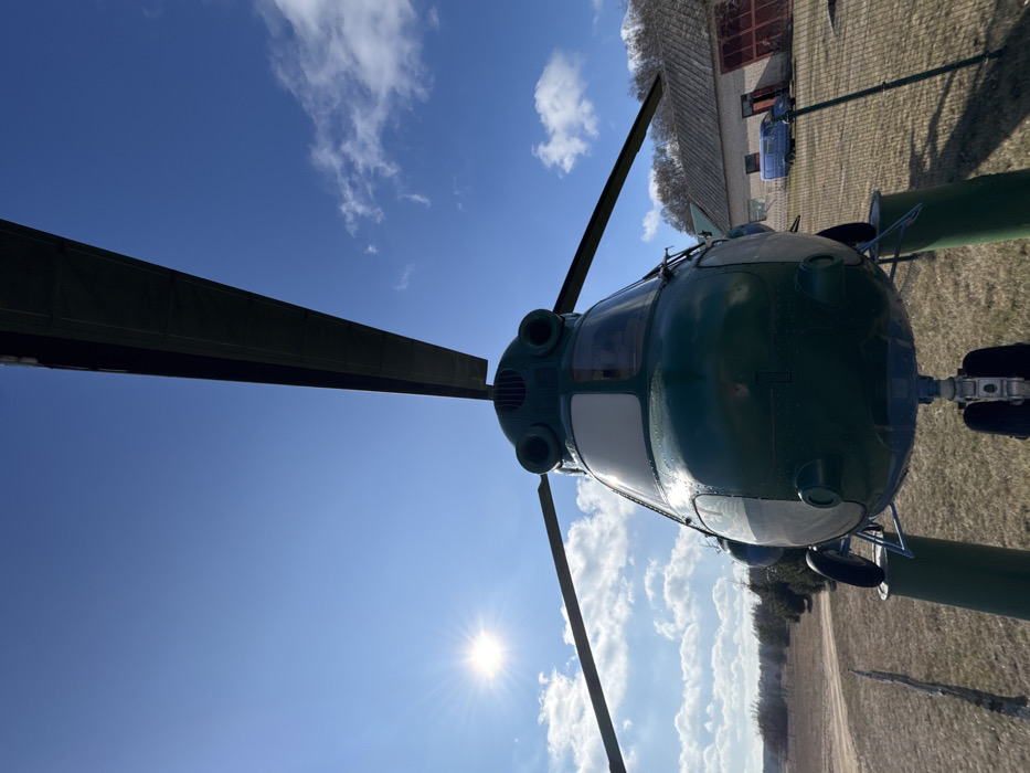 A large, dark green Mil Mi-2 type helicopter, featuring prominent main rotor blades and a visible cockpit, occupies the central foreground of the image. The helicopter is static on a patch of dry, light brown grass, with its landing gear visible. The scene is set under a bright blue sky with scattered white cumulus clouds. The sun is intensely bright in the upper left quadrant, creating a starburst effect. To the left of the helicopter, an open field of dry grass extends towards a distant line of dark evergreen trees. To the right, a single-story brick building with a dark sloped roof and two large, dark red doors is visible. A light blue utility van is parked in front of this building. No individuals are present. This scene is located in Падлабенскі сельскі Савет, Belarus.
