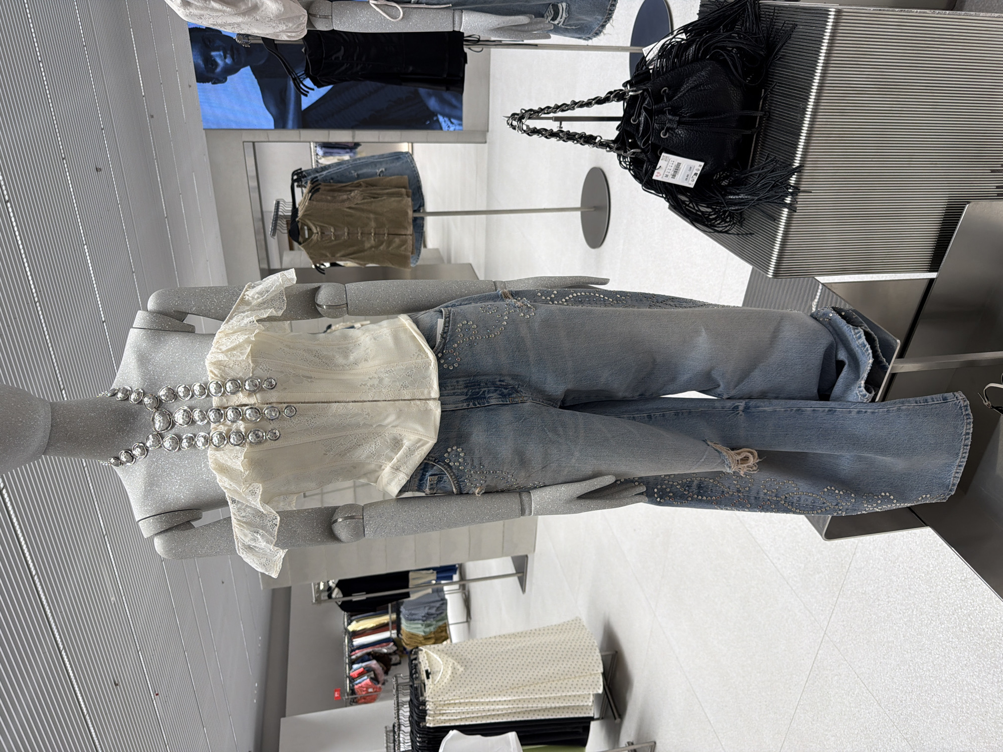 An off-the-shoulder white lace corset with a ruffled neckline is displayed on a grey mannequin in a store. The mannequin wears light blue wide-leg jeans embellished with rhinestones and featuring a ripped section. A chunky silver necklace adorns the mannequin's neck. To the right, a black fringe bucket bag with a chain strap is placed on a ribbed metal pedestal. In the background, racks of clothing and a large digital screen displaying a person's portrait are visible. The floor is a light-colored tiled surface, and the ceiling has linear lighting.