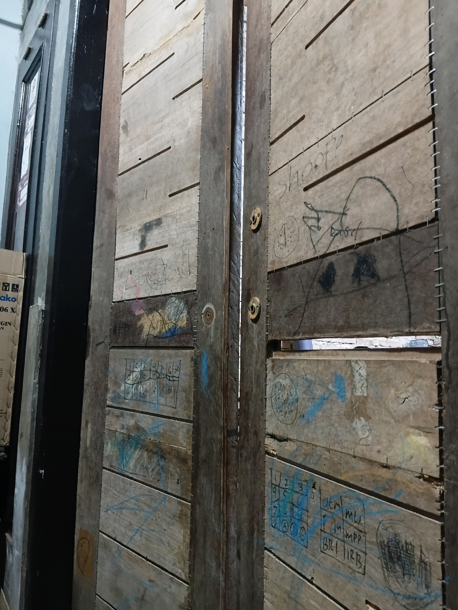 The image shows a close-up view of a wooden door with various markings and scribbles on its surface. The door appears to be made of horizontally oriented wooden planks, with visible stitching or fasteners along the seams. The wood has a weathered and aged appearance.
On the left side of the door, there is a vertical dark structure, possibly another door or frame, with a reflective surface. To the left of that, a sliver of a white object with some text and graphics is visible, resembling a cardboard box or packaging. The text is partially legible, with "ako" and "06 X" visible.
The main focus is the weathered wooden door. The surface of the door is covered with a variety of markings. These include:
* **Text:** Several instances of handwritten text are visible. Some appear to be words like "shoopp" and what looks like "BRI IRBB". Other text is illegible or fragmented.
* **Drawings/Symbols:** There are various drawings and symbols. These include what looks like a crudely drawn face or head outline, some circular patterns, and abstract shapes.
* **Numbers:** A grid of numbers, resembling a keypad or a game board, is drawn on one of the planks. The numbers range from 1 to 9, with some symbols like circles and what might be a letter "O" or a smiley face.
* **Color:** Some of the markings are made with colored chalk or crayon, with traces of blue, yellow, and pink visible. There are also black markings, likely from a pen or marker.
The door has two circular metal fixtures, possibly doorknobs or decorative elements, on its vertical edge. The lighting in the image is dim, casting shadows and highlighting the texture of the wood.
The image is taken from a low-angle perspective, looking upwards at the door. The scene suggests an outdoor or semi-outdoor environment, possibly a storage area or an alleyway, given the weathered materials and the nature of the markings. The specific location in Cikal, Indonesia, is not directly evident from the visual cues within the image itself, but the request implies this context.