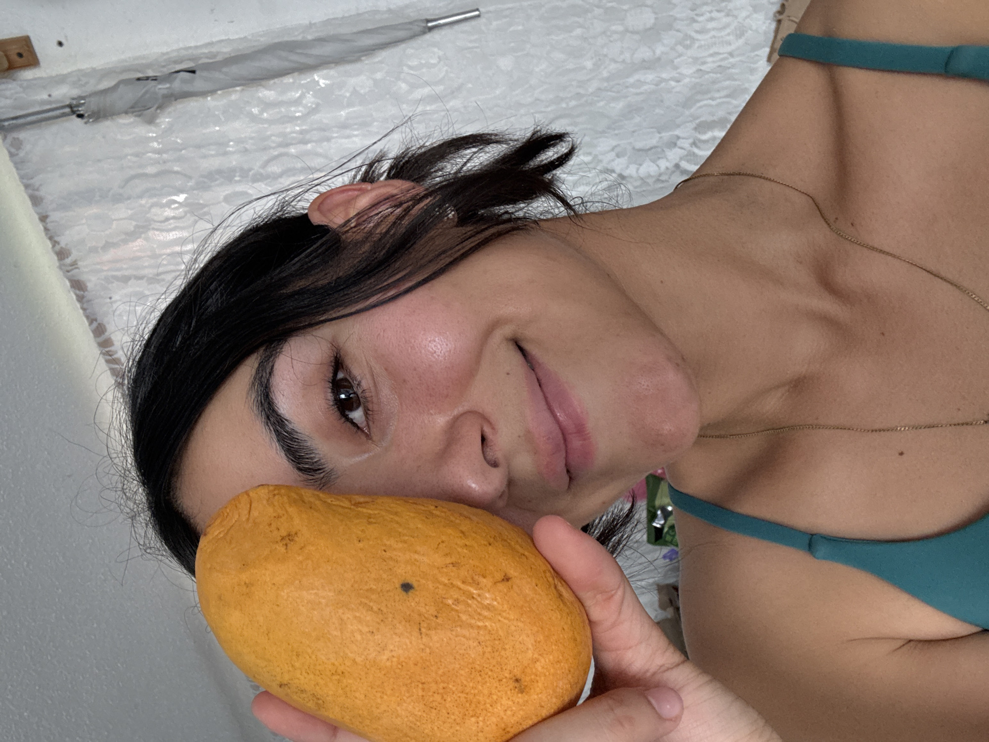 A dark-haired woman with fair skin holds a mango in front of her face. She wears a green top and a gold chain necklace. The background includes a white wall, an umbrella, and a white patterned surface. The location is likely indoors, but there is no other indication of Puebla, Mexico.