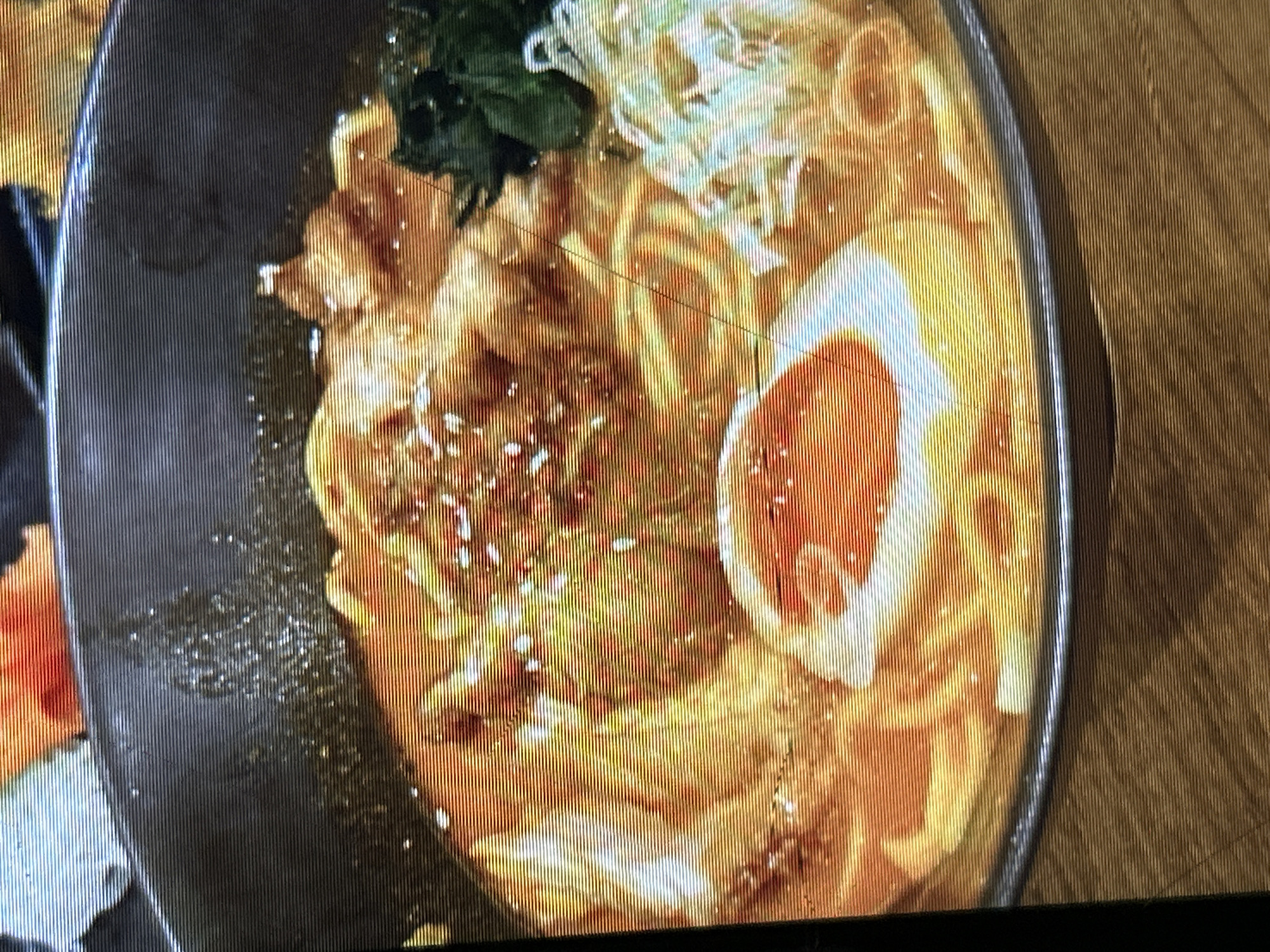 The image shows a close-up of a bowl of ramen noodles. The ramen is in a dark bowl, and the broth appears to be a reddish-orange color. The noodles are yellow and thin. There is a soft-boiled egg cut in half, with the yolk exposed, as well as some pieces of meat and green vegetables. The background is blurred, but it appears to be a wooden table. There are no people or any other identifiable objects in the image besides the food. The image appears to be taken in a restaurant setting. There is no information in the image to suggest the location is Depok, Indonesia.