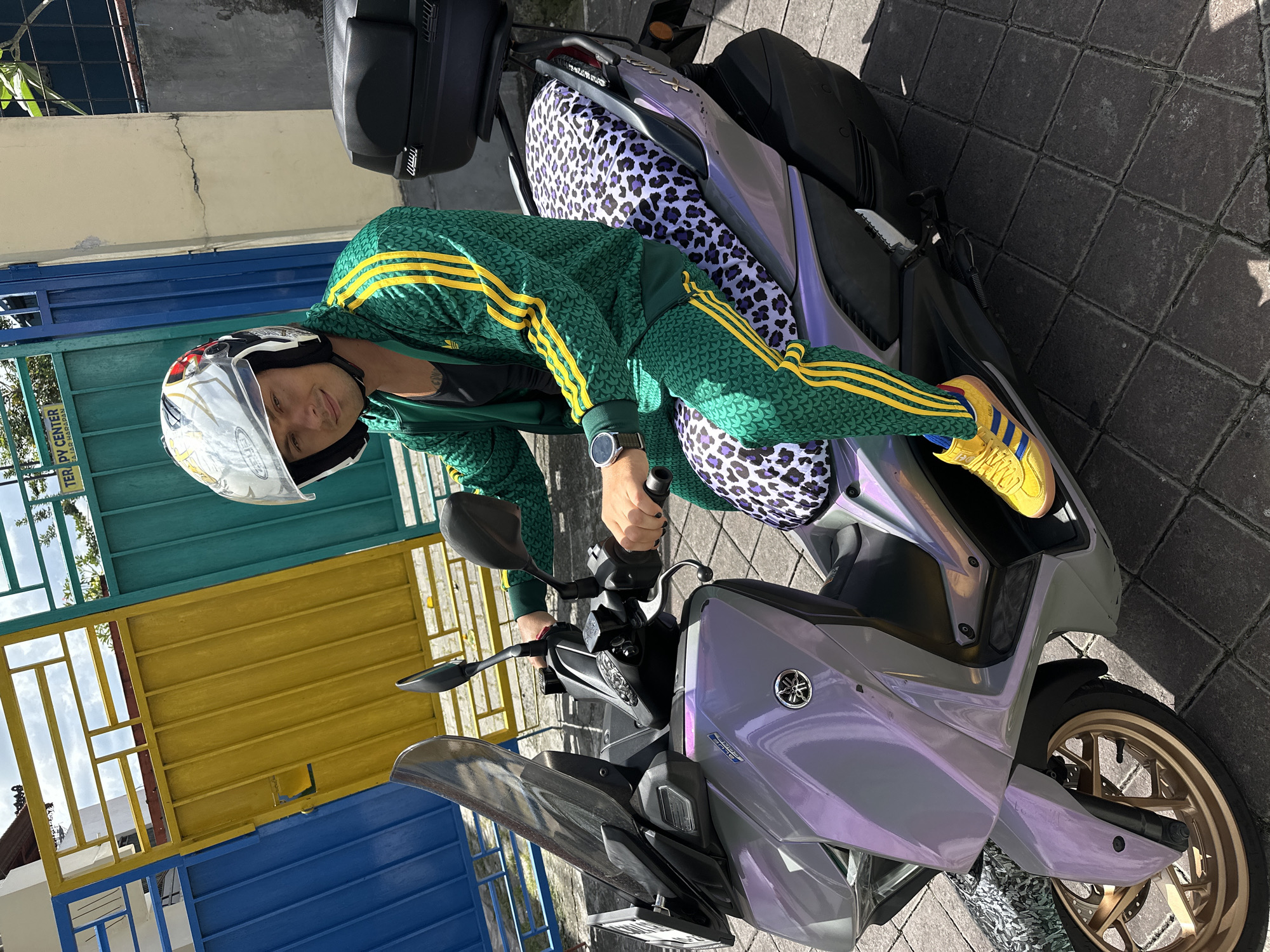 A man in a green and yellow tracksuit, wearing a white helmet, is sitting on a silver-purple Yamaha motorcycle with a leopard print seat. He is smiling at the camera. The motorcycle is parked on a stone-paved surface in front of a gate painted in turquoise, yellow, and blue. The location is North Kuta, Indonesia.