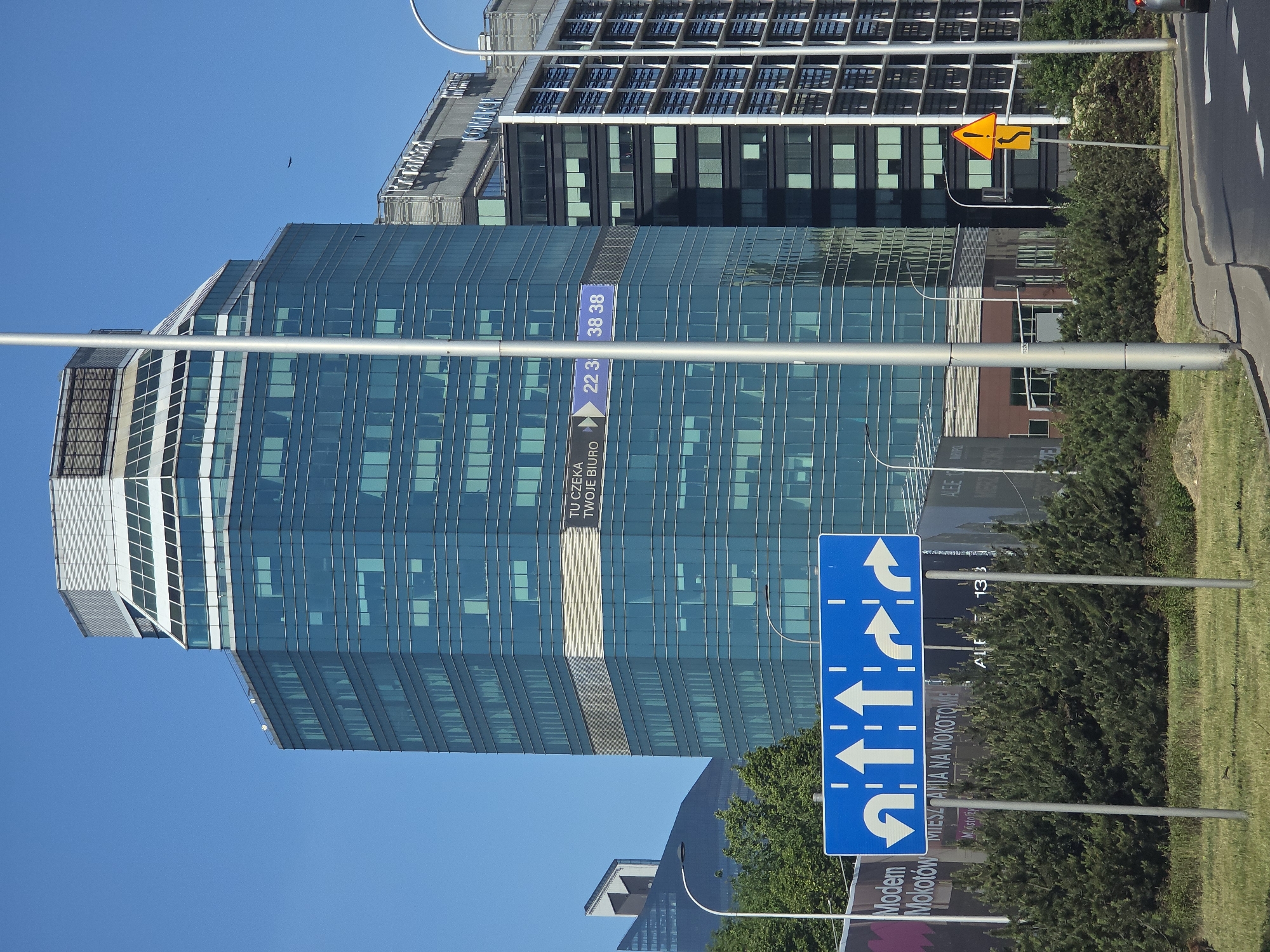 A medium, eye-level shot, rotated clockwise, shows a street scene in Warsaw, Poland with modern architecture and road signage under a clear blue sky.
Dominating the frame is a modern, glass-clad skyscraper, its surface reflecting the sky and other buildings. A vertical band runs across the building’s façade with text in Polish "TU CZEKA TWOJE BIURO", accompanied by the numbers "22 3" and "38 38." Another building is visible behind the skyscraper.
To the right of the skyscraper, a yellow traffic sign with a snake-like curve symbol stands at the edge of the road. A large, blue traffic sign in the foreground indicates the available lanes for traffic. It shows various arrows pointing straight, left, and for U-turns, with text "MIESZANIA NA MOKOTOW" and "Modern Mokotów". Street lights and trees are interspersed throughout the scene. The road is partially visible, with a light-colored sidewalk to its right.