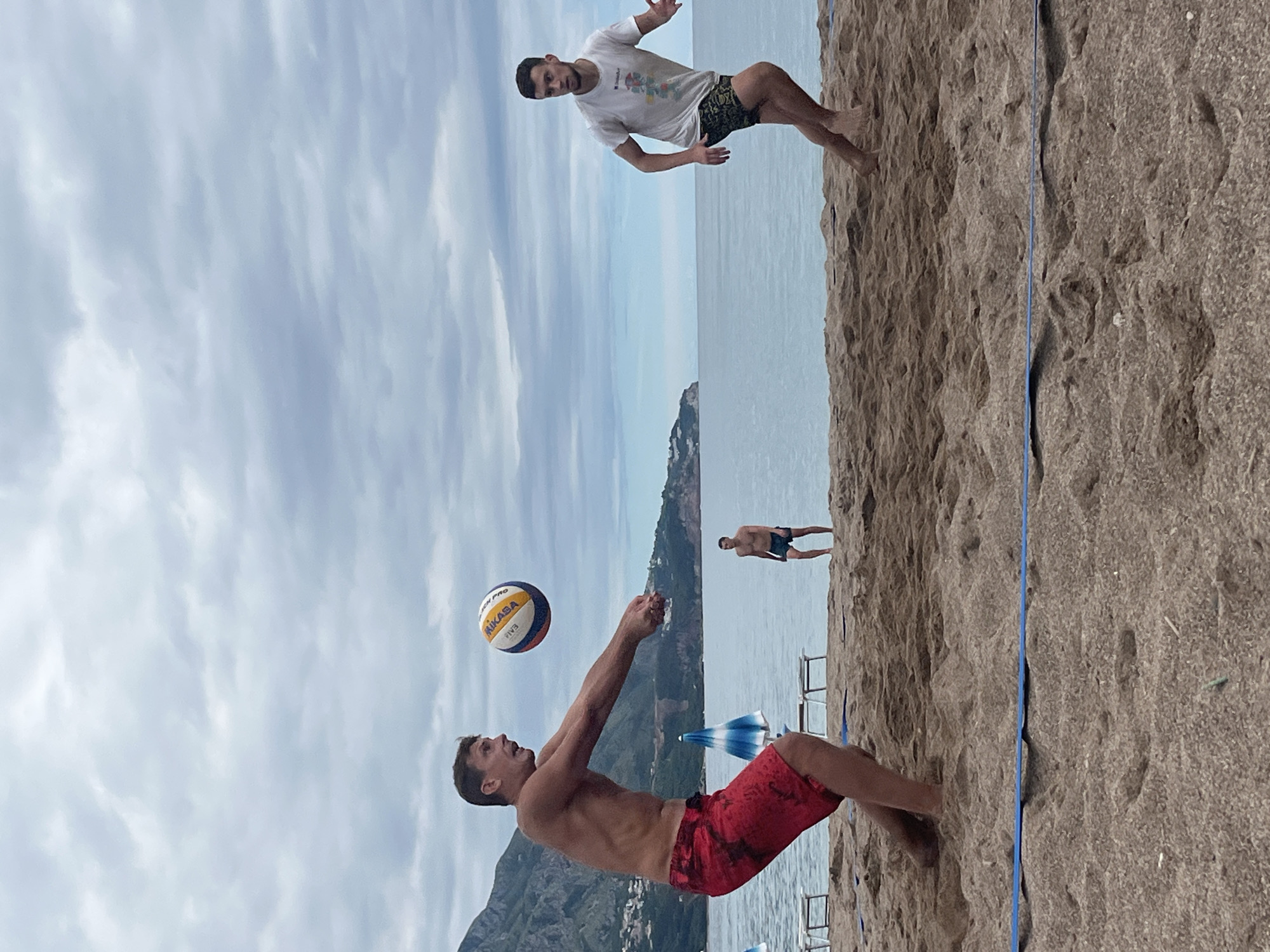 The image captures a beach volleyball scene in Boreti, Montenegro. In the foreground, a shirtless man in red swim trunks bumps a volleyball, presumably during a game. Behind him and slightly above, another man in a white t-shirt jumps, anticipating the ball. In the background, a third man stands in the water near the shore. The sea and mountains are visible in the background, along with beach umbrellas and chairs. The sky is cloudy. A blue volleyball net runs across the sand.