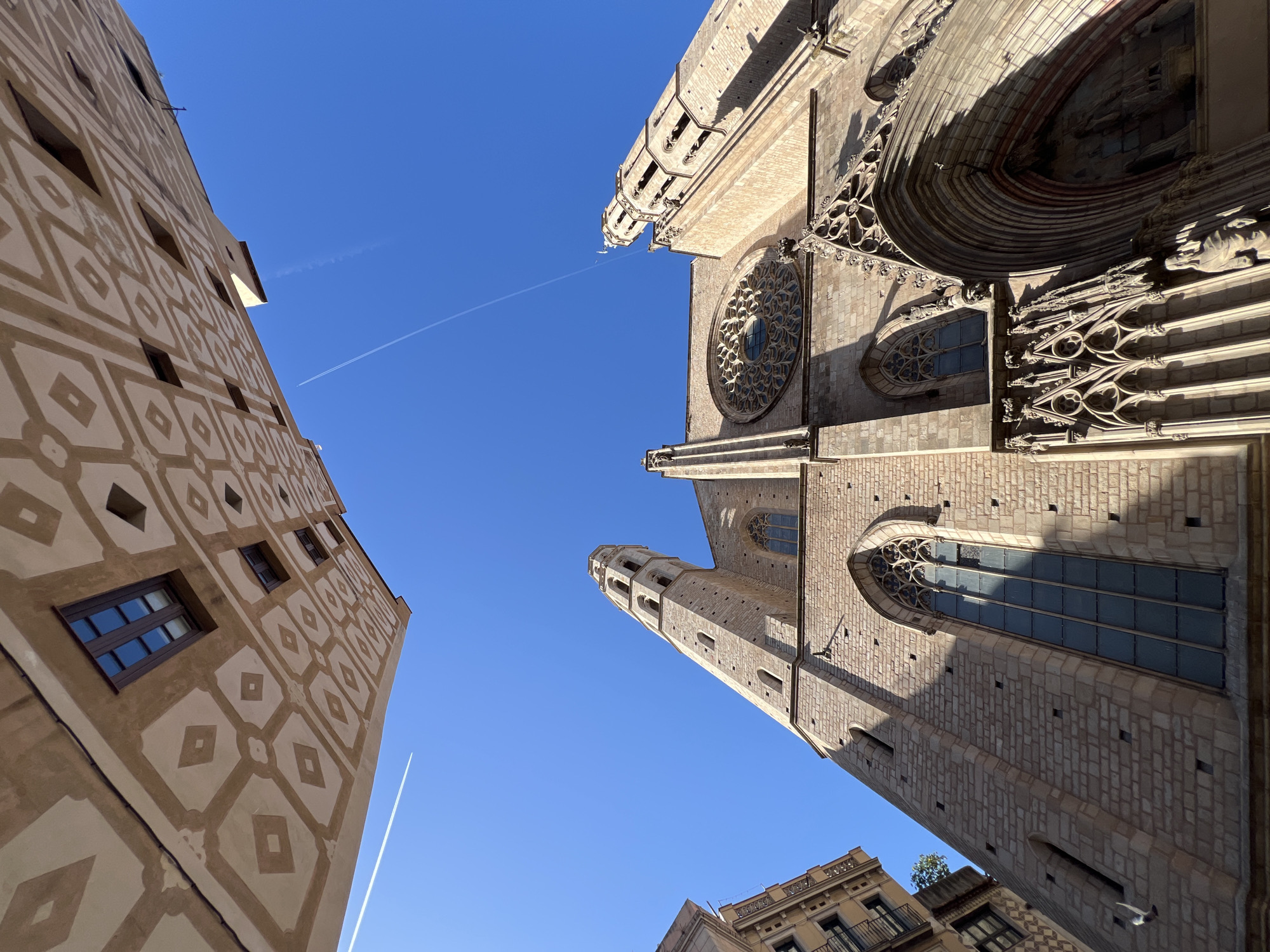 This low-angle photograph captures the upper facade and bell tower of the Basilica of Santa Maria del Mar in Barcelona, Spain. The Gothic basilica, constructed from light-colored stone, features a prominent circular rose window with intricate tracery on its main facade, flanked by a tall, octagonal bell tower on the right. Below the rose window, tall, arched windows with clear glass are visible. A section of an ornate entrance portal with detailed carvings and statuary is partially visible at the bottom right. Direct sunlight illuminates the church's stone surfaces, casting defined shadows. In the upper left quadrant, a portion of an adjacent building's facade is visible, characterized by a light-brown color adorned with a repeating pattern of diamond and geometric shapes, incorporating two dark-paned windows. The upper half of the image is dominated by a clear, deep blue sky, traversed by two thin, white airplane contrails.