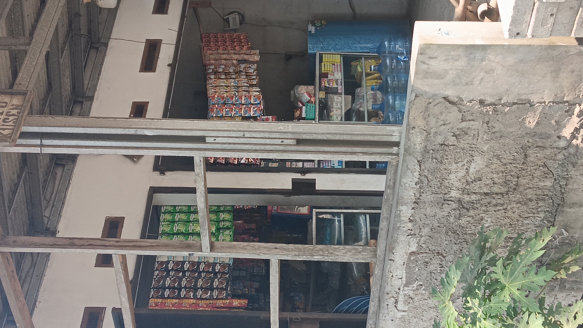 This is a full, eye-level shot of a small, open-air shop in Jonggol, Indonesia. The shop appears to be a convenience store or warung selling a variety of goods, including snacks, bottled water, and possibly household items. The shop is constructed with a metal roof and white walls. In the foreground, a rough concrete wall with some greenery in the lower left corner separates the viewer from the shop.
Inside the shop, there are several shelves displaying various products. Packages of snacks are hanging from the ceiling in neat rows. On a shelf behind the counter, there are several large bottles of drinking water. A blue refrigerator stands to the right of the shelves. The shop appears to be open for business, though no customers or staff are visible in the frame. The overall impression is of a typical, modest neighborhood store. The sign above the shop, partially visible, appears to have text in Indonesian.