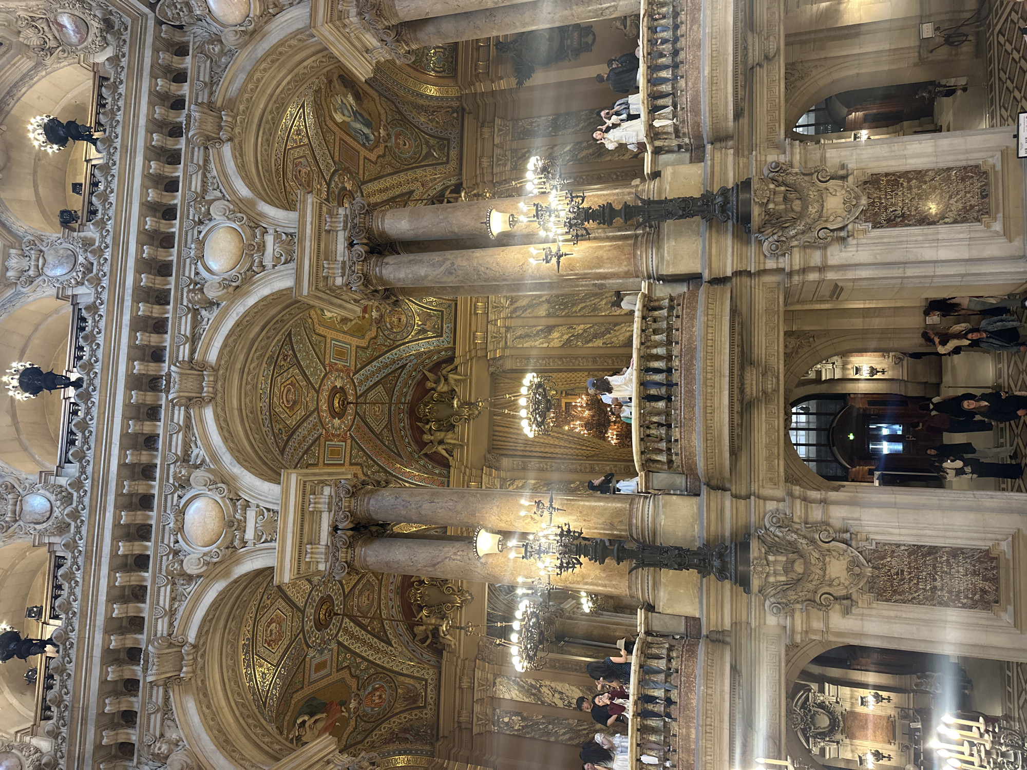 This image presents the opulent interior of a grand building, likely a historical landmark, with multiple levels and intricate architectural details. The scene is dominated by classical-style architecture featuring soaring marble columns, richly decorated vaulted ceilings, and ornate arches. The ceiling and upper wall sections are adorned with gilded mosaics, frescoes depicting figures, and complex geometric patterns.
Numerous people are scattered throughout the scene, appearing to be visitors or tourists. Some are visible on the lower level, while others lean over the elaborate balustrades of balconies, observing the grand space. They are dressed in casual, modern attire.
The setting is illuminated by large, multi-tiered chandeliers with electric lights, complemented by smaller wall sconces, casting a warm glow throughout the area. The overall impression is one of immense scale and luxury.
A notable detail is a dark marble memorial plaque mounted on a prominent pillar. Partially legible text on the plaque includes the French phrases "MORTS POUR LA FRANCE" (Dead for France) and the date range "1914-1918," indicating a World War I memorial. Further text reads "AUX MEMBRES DU PERSONNEL DECEDES AU CHAMP D'HONNEUR" (To the members of the staff/personnel deceased on the field of honor).
The activity centers around observation and appreciation of the architectural beauty. The time of day appears to be daytime, with artificial lights enhancing the interior's splendor. Given the context of Paris, France, the architectural style and memorial suggest a significant public building, such as the Palais Garnier (Paris Opera House).