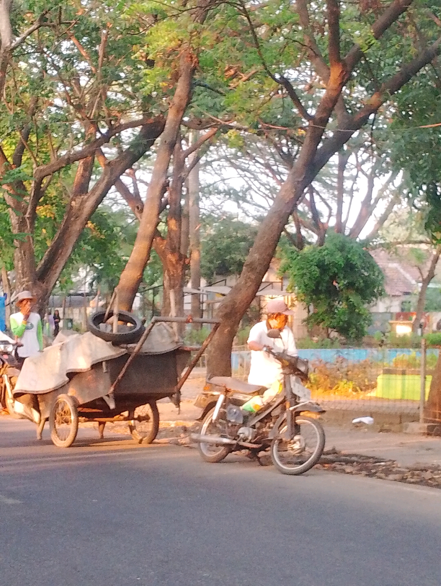 In Probolinggo, Indonesia, two individuals are depicted outdoors near a street. On the left, a person wearing a hat and a light-colored shirt with a green sleeve is standing next to a cart. The cart, which has two large wheels and appears to be laden with covered goods, is also topped with a spare tire. To the right of the cart, another individual is seated on a motorcycle. This person is wearing a white shirt and a hat. The background shows lush green trees with visible branches, a weathered wall, and a hint of buildings with red tile roofs. The ground is paved, and sunlight casts a warm glow on the scene, suggesting it is either early morning or late afternoon.