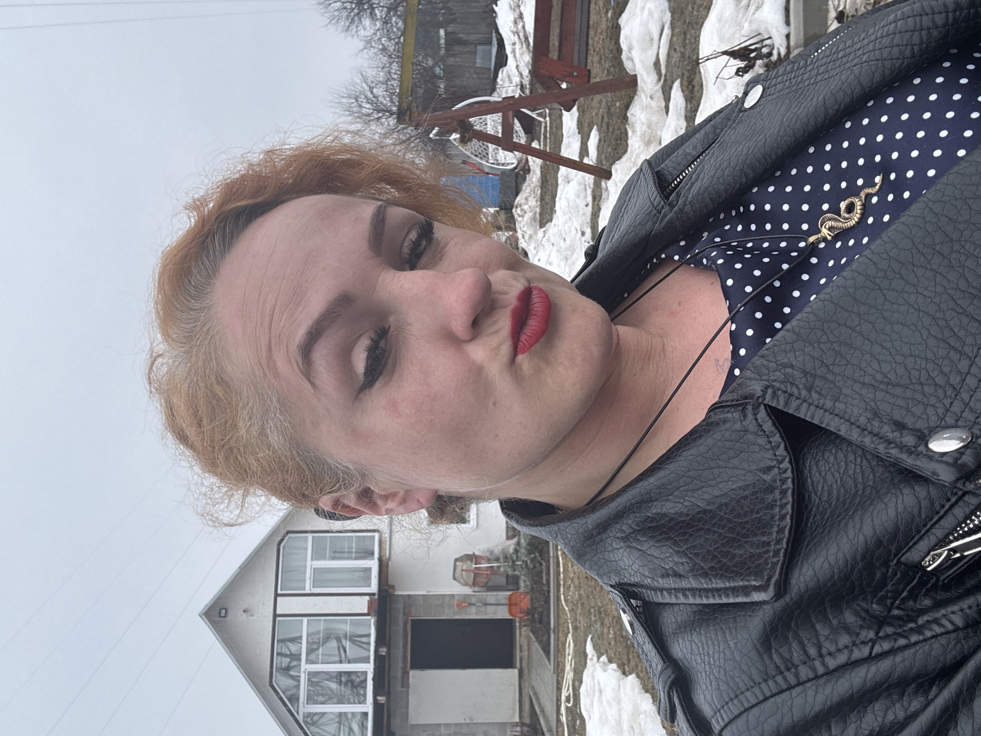 The image depicts a woman with auburn hair, red lipstick, and visible eyelashes, wearing a black leather jacket over a navy blue polka dot dress with a gold snake pendant necklace. She is outside, likely in a backyard or residential area, as evidenced by a house in the background with white siding and multiple windows. Patches of snow are visible on the ground, and there is a wooden structure behind her, possibly a swing set or some type of outdoor furniture. It is a cloudy day, indicated by the flat, overcast sky. The woman is looking directly at the camera with a slight pucker of her lips. The location is the city of Chaikovsky, Russia.