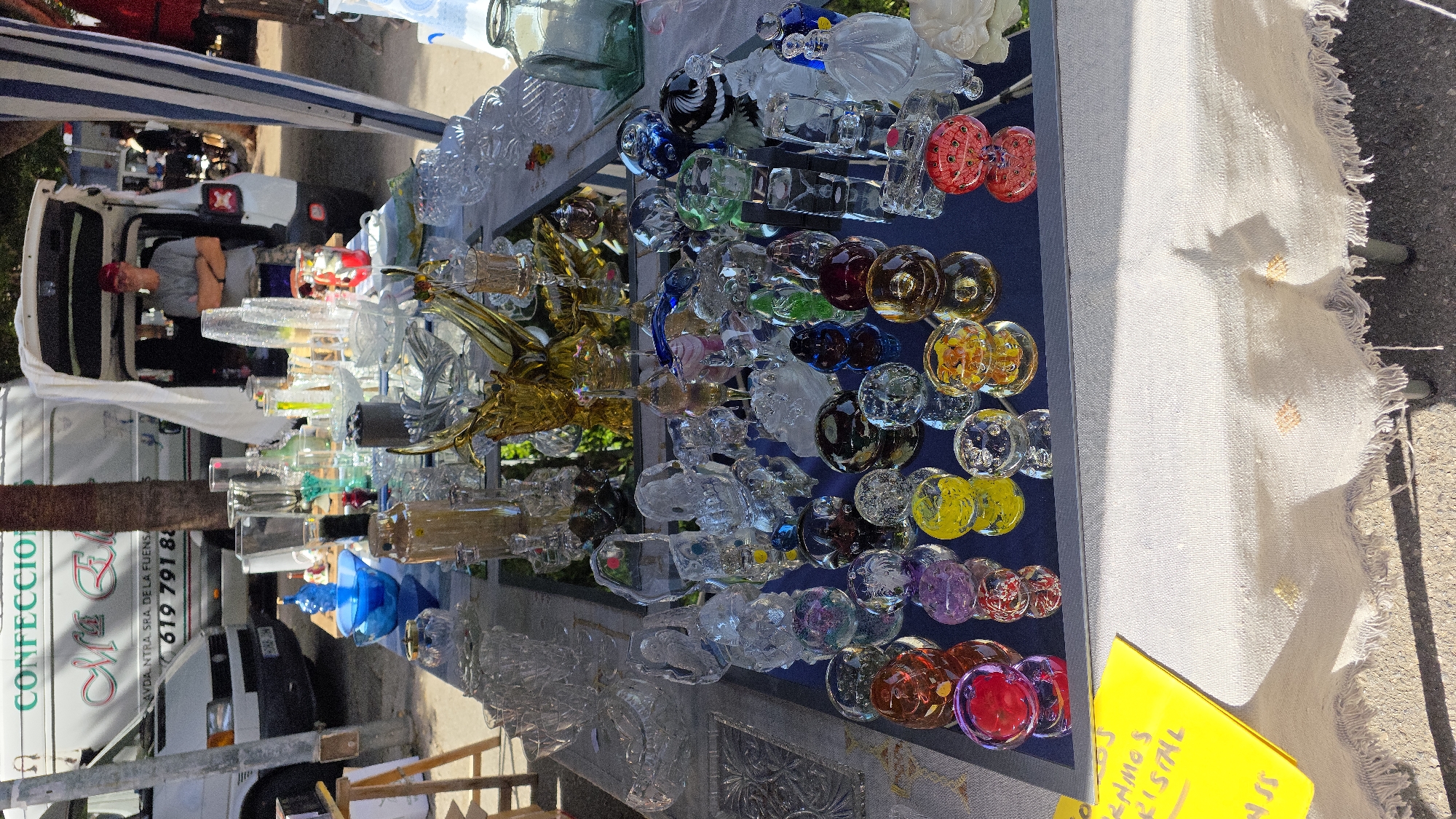 A high-angle medium shot shows a street vendor's table laden with a variety of glassware, displayed outdoors. The table is covered with a white cloth that has frayed edges and is adorned with embroidered yellow and orange accents. To the left, a bright yellow sign with handwritten text in Spanish reads "OFRECEMOS CRISTAL" ("We offer crystal"). Behind the table, a white van with Spanish text and a phone number "619 791 88" is visible, suggesting the vendor might be operating out of it. A man wearing a red baseball cap and a grey t-shirt stands with his arms crossed behind the open back of a white vehicle, possibly a Jeep, which is parked directly behind the table. The scene is set in Almuñécar, Spain, as indicated by the presence of Spanish text and the general appearance of a street market environment. The background reveals trees and other market stalls, suggesting a bustling outdoor market. The glassware on display includes a diverse assortment of clear and colored glass items, such as bowls, vases, figurines, and decorative spheres.