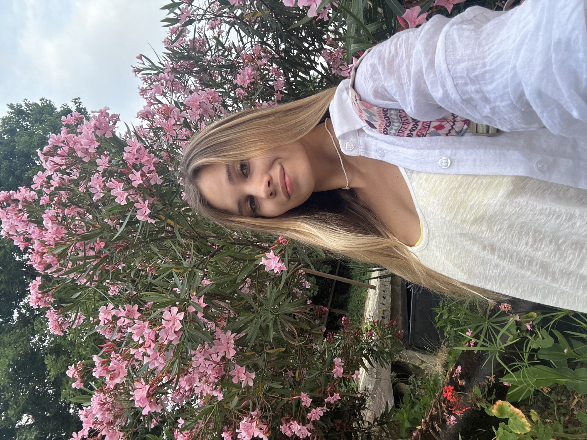 A young woman with blonde hair is taking a selfie in front of a bush with pink oleander flowers. She is wearing a white linen shirt over a yellow tank top. Her left arm is extended to capture the photo. The background shows lush green foliage and a cloudy sky. The scene appears to be outdoors, likely in a garden or park. The image does not contain any information that places it specifically in Sokhumi, Abkhazia.