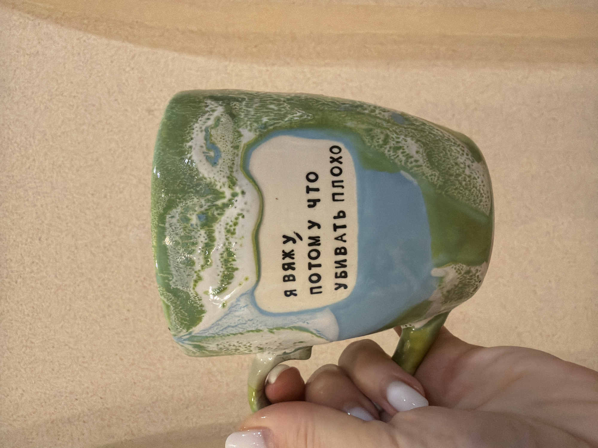 A hand holds a green and blue glazed ceramic mug with the Russian text "Я вяжу, потому что убивать плохо" ("I knit, because killing is bad"). The background appears to be a light beige wall. There is no information to definitively place this image in Levkovo, Russia.