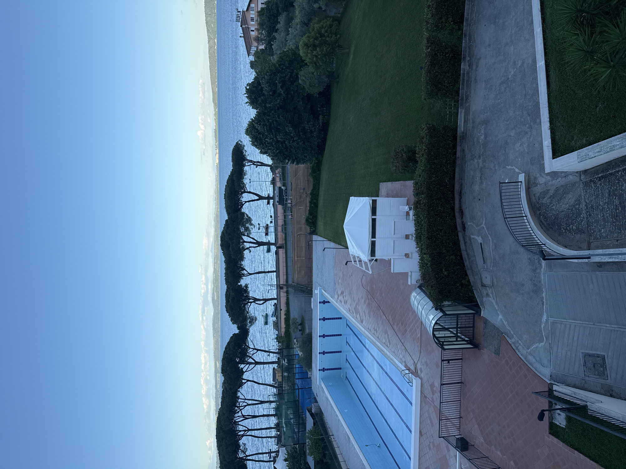 The image shows an elevated view of a swimming pool complex situated near Lake Bracciano in Italy. The pool is rectangular with multiple lanes and blue water. Adjacent to the pool is a small, white structure and a partially enclosed walkway. A line of tall, thin trees separates the pool area from the lake, which stretches into the distance under a clear blue sky. Further along the shoreline, a building is visible amidst the trees. A well-maintained green lawn borders the pool on one side. To the right, a paved pathway with railings and a small patch of green grass are discernible, suggesting this is an area for movement. The lighting suggests either early morning or late afternoon.