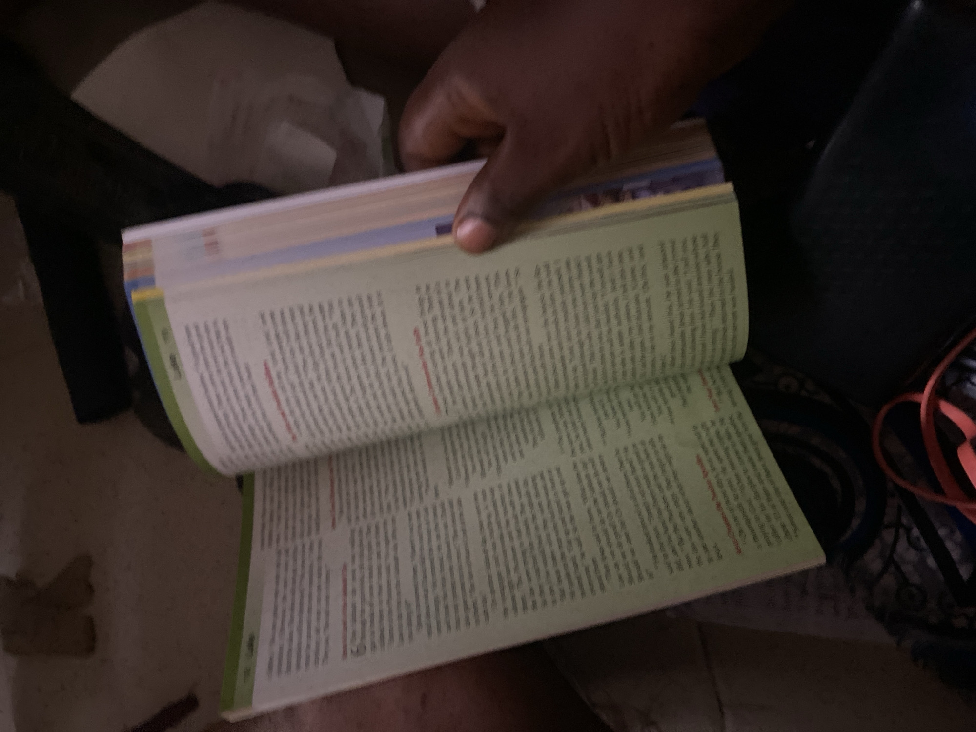 The image shows a close-up of an open book with text on its pages. A hand, appearing to be of a person of color, is holding the book open and turning a page. The book has a green section on the top edge and colored tabs on its side. The background is dark and out of focus, with some objects visible but indistinct. The image is taken from a high angle, looking down at the book. There is no identifiable information within the image to suggest a location in the city of Jauro Yinu, Nigeria.
