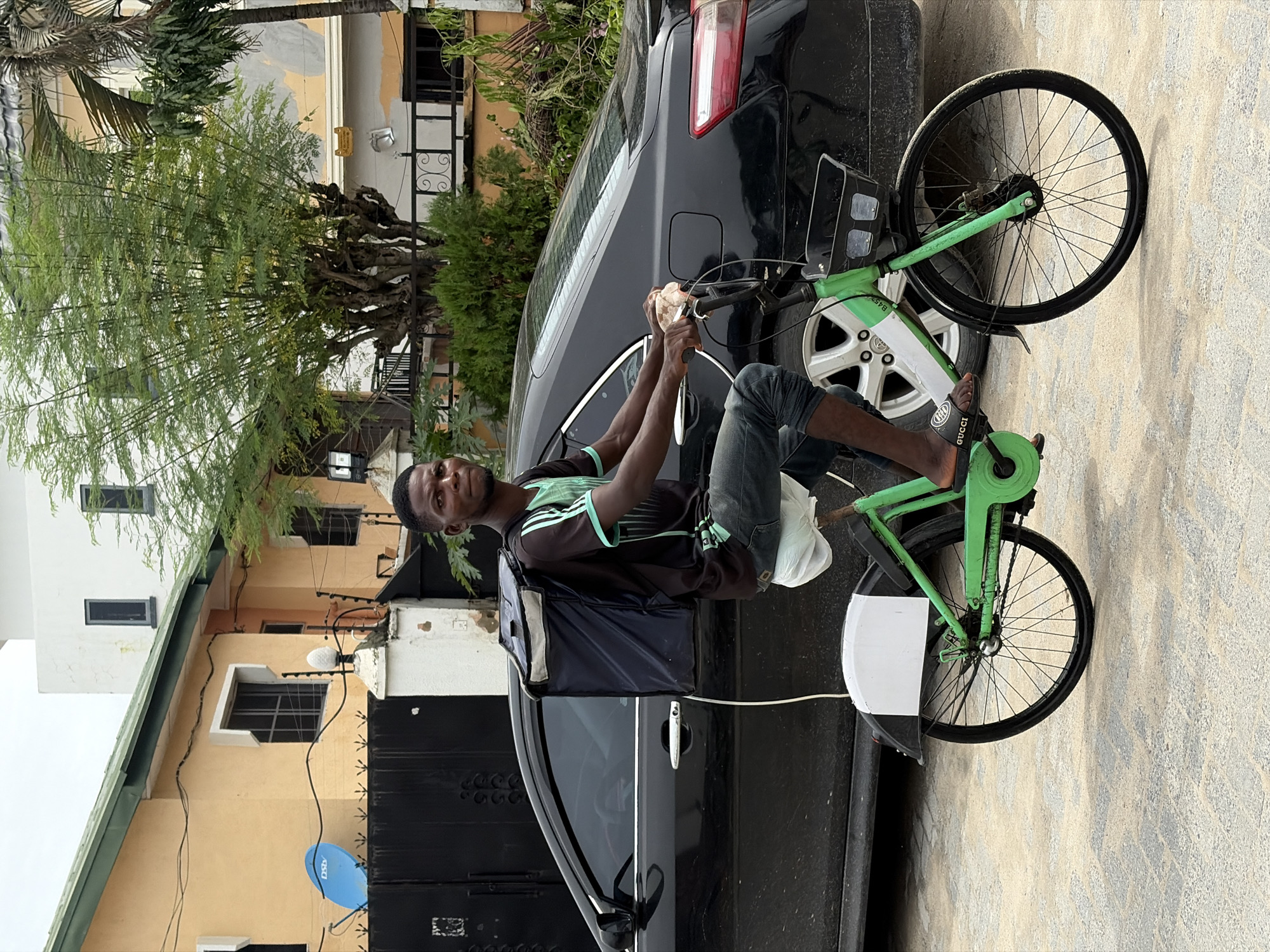 A man wearing dark jeans, a dark t-shirt with light green stripes, and Gucci sandals is seated on a green bicycle in Igboefon, Nigeria. He has a dark-colored backpack and holds a white bag. The bicycle is parked next to a black car. The background features buildings, trees, and a satellite dish. The image has been rotated 90 degrees clockwise.