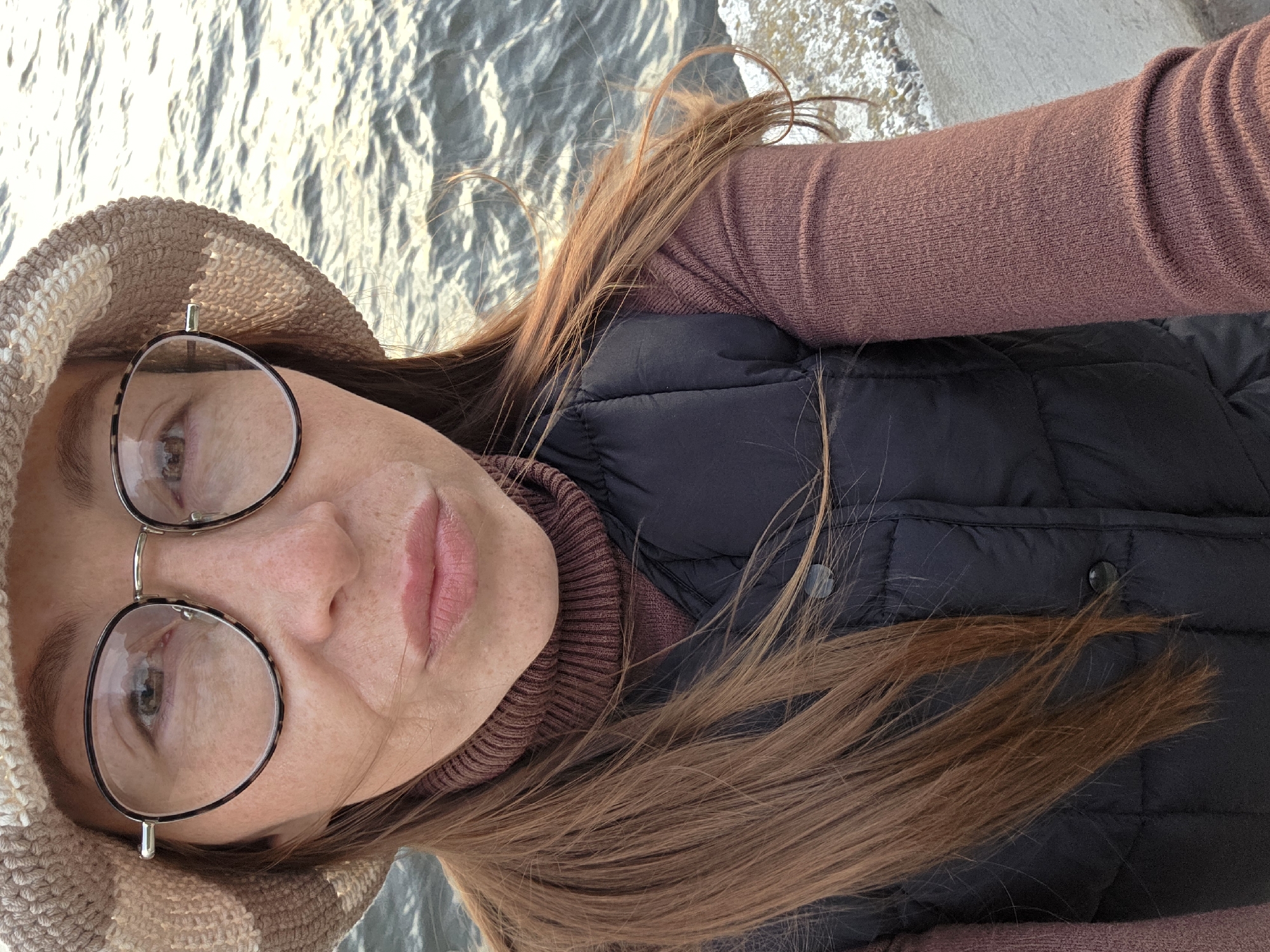 A person with long, brown hair, wearing a brown turtleneck sweater and a black puffer vest, is posing for a selfie. They are wearing round-framed glasses and a beige and white striped hat. The background appears to be a body of water, possibly a river or lake, with sunlight reflecting on its surface. Based on additional information about the location, it is presumed that this scene is located in the city of Chaikovsky, Russia.